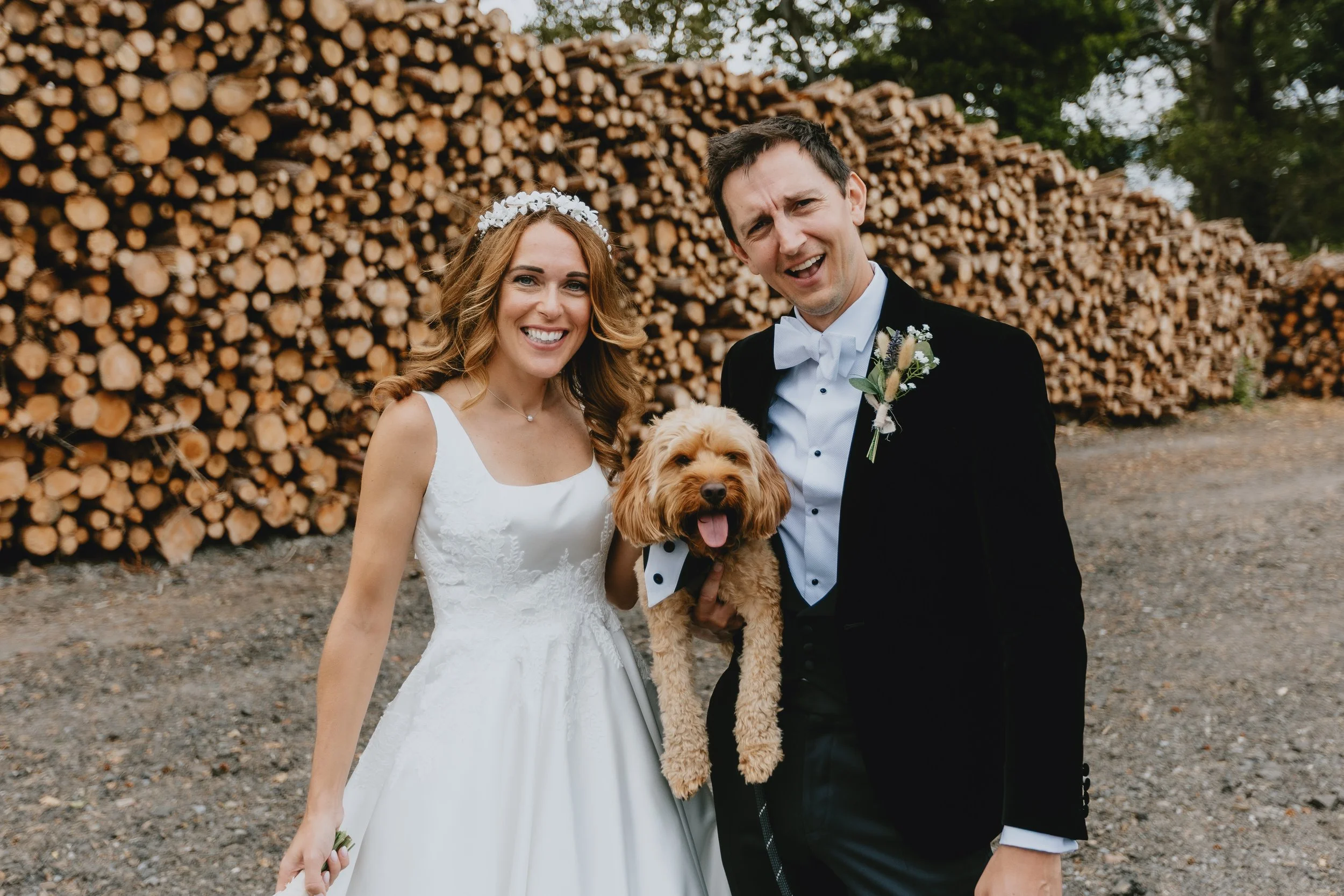 A happy bride and groom stand outdoors with a dog in front of stacked logs. The bride wears a white wedding dress and a floral headband, while the groom wears a black tuxedo with a white bow tie and boutonniere. Both are smiling widely.