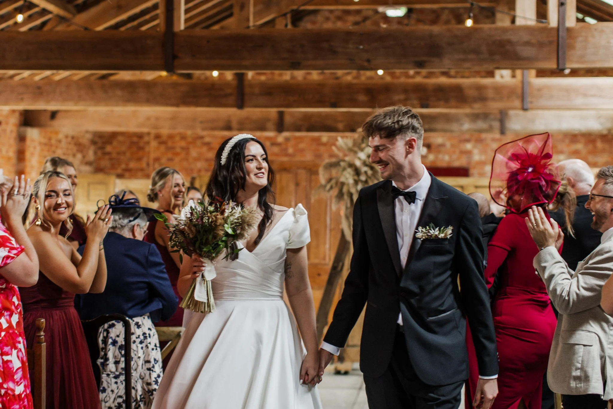 A bride and groom holding hands and smiling during their wedding ceremony with guests clapping in the background.