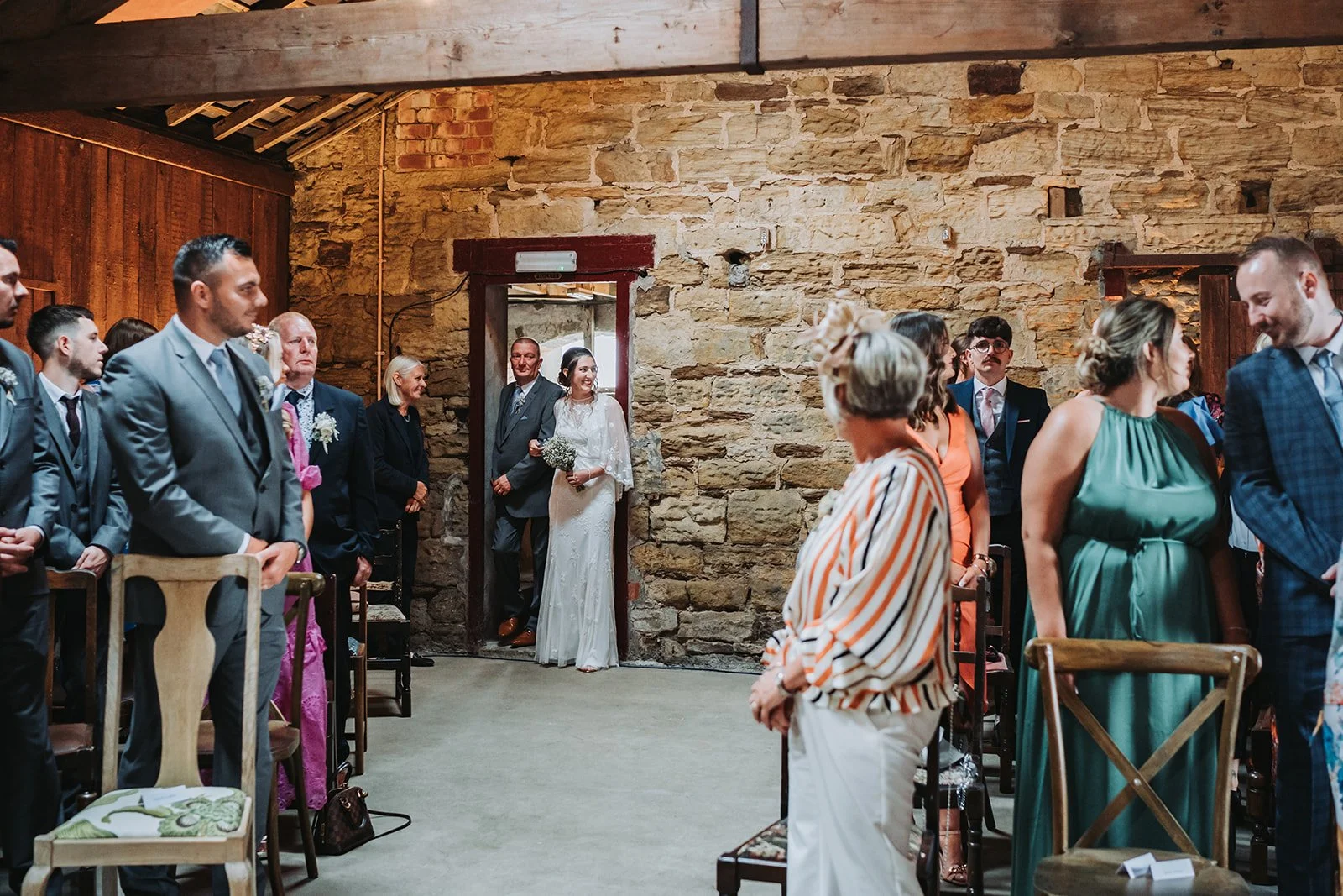 A bride and a man, likely her father, walking into a wedding ceremony in a rustic stone-walled venue filled with guests, some standing and some sitting, dressed in formal attire.