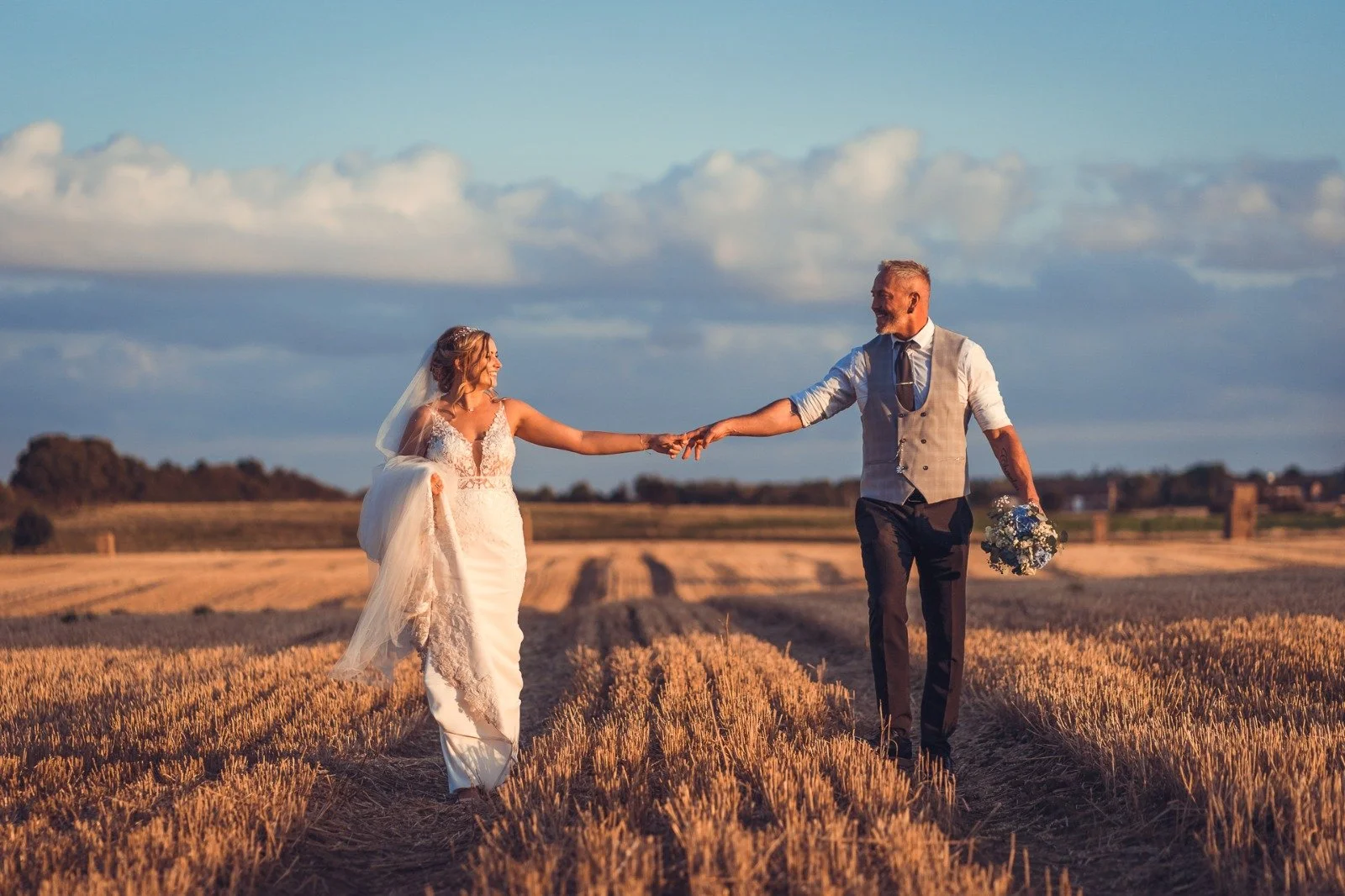 Bride and groom holding hands and walking through a wheat field at sunset, with a cloudy sky in the background.