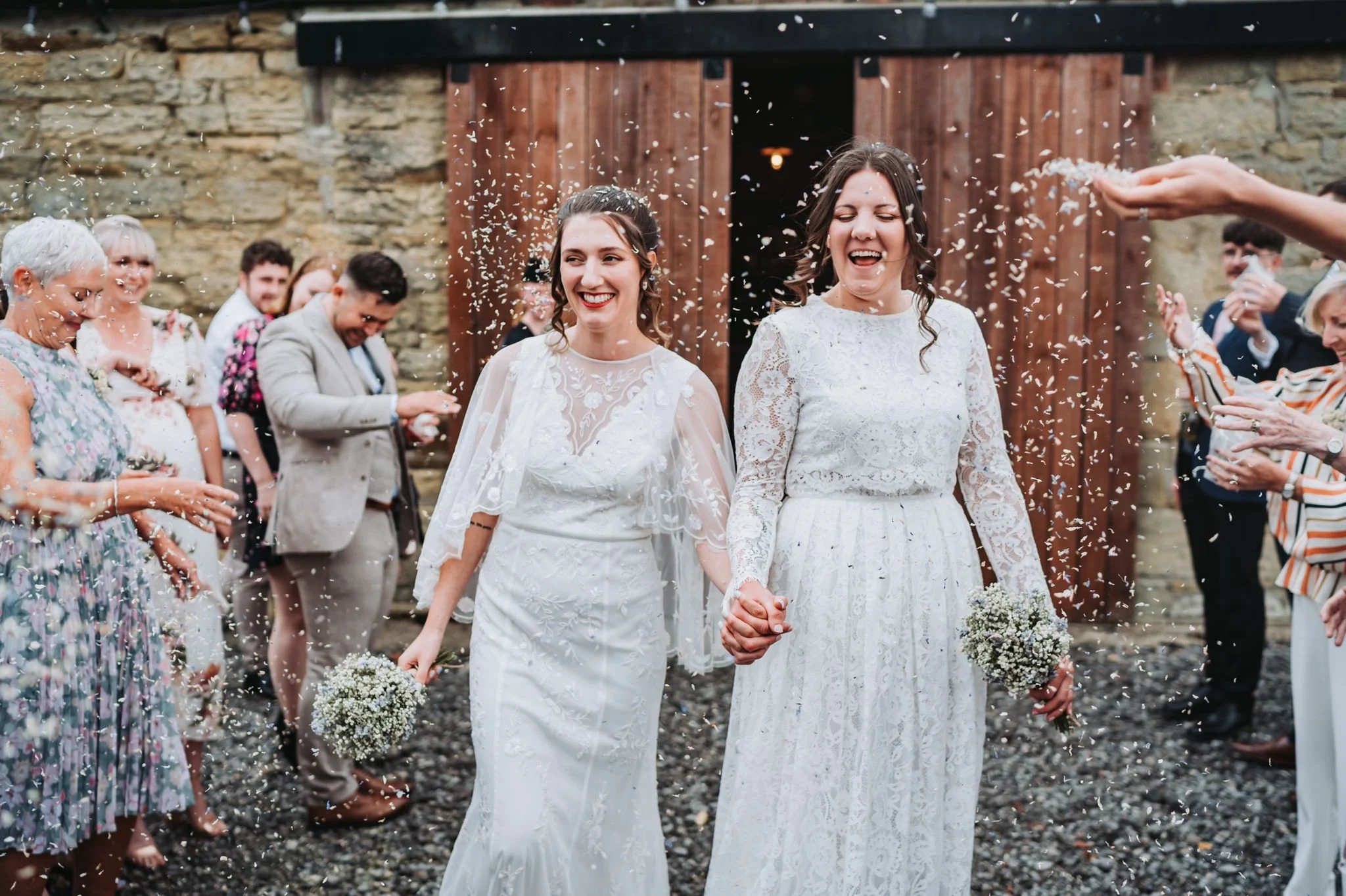 Two women in white wedding dresses holding hands and smiling, surrounded by friends and family throwing rice outdoors.