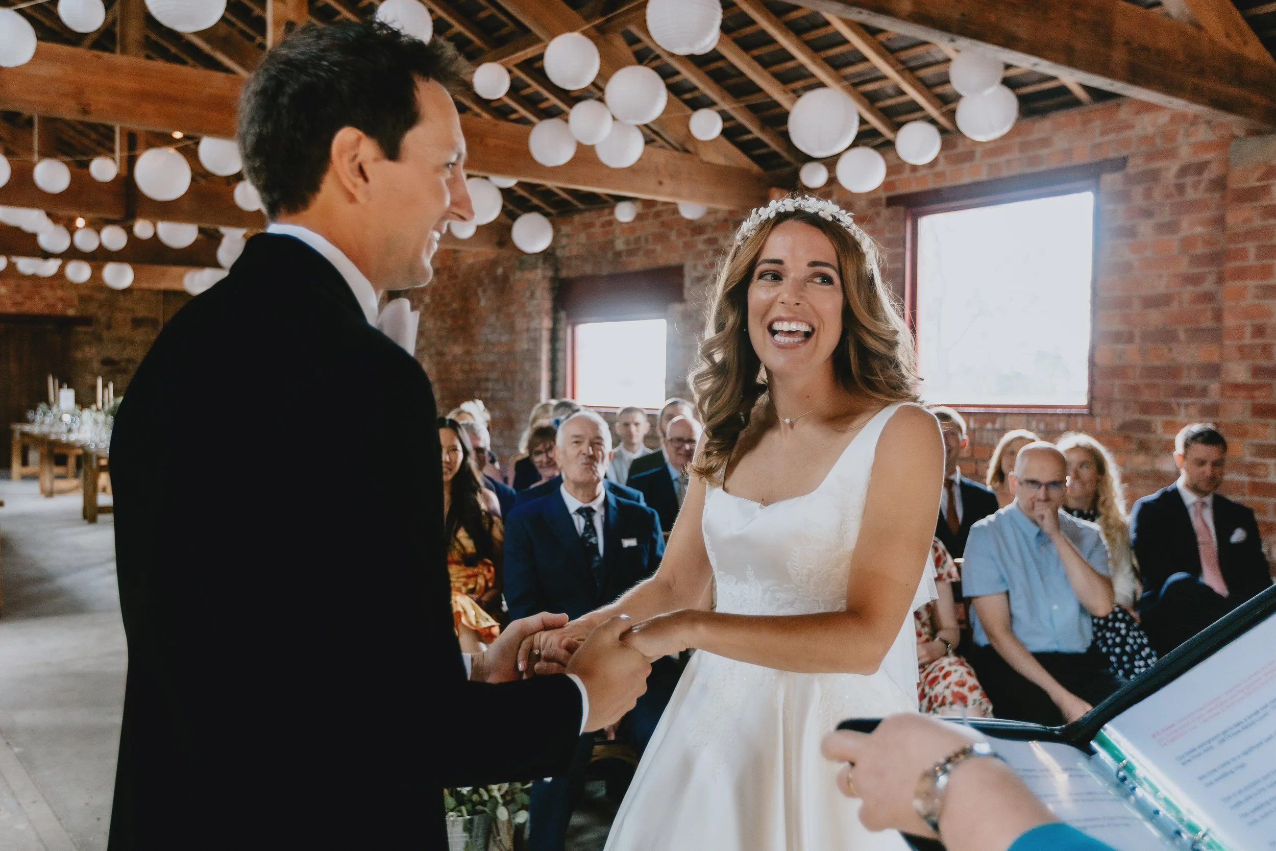 A couple exchanges vows during a wedding ceremony in a rustic barn with string lights and exposed brick walls, with guests seated in the background.