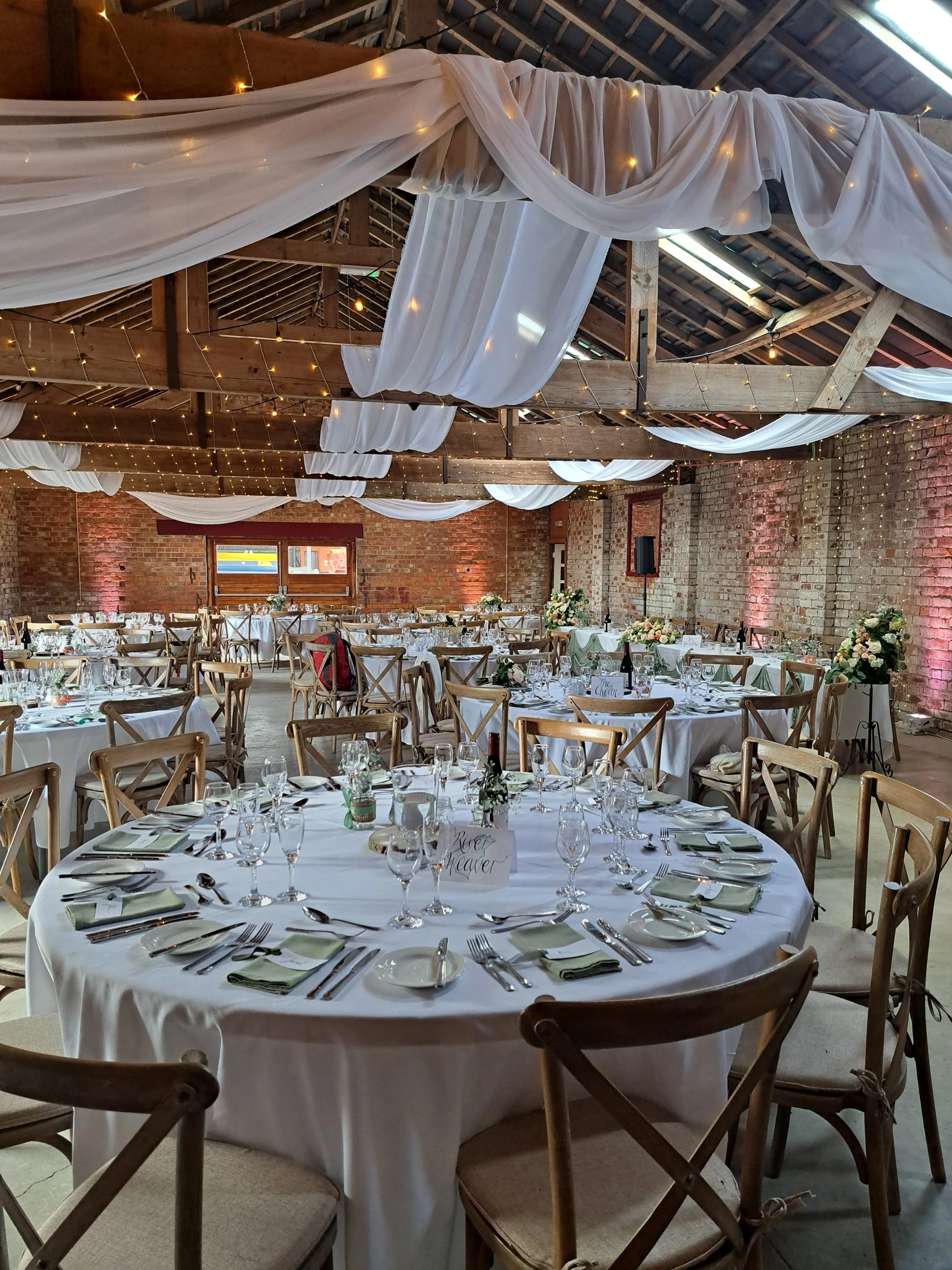 Wedding reception setup in a rustic barn with string lights, white draped fabric, and decorated round tables with white tablecloths, glasses, and floral centerpieces.