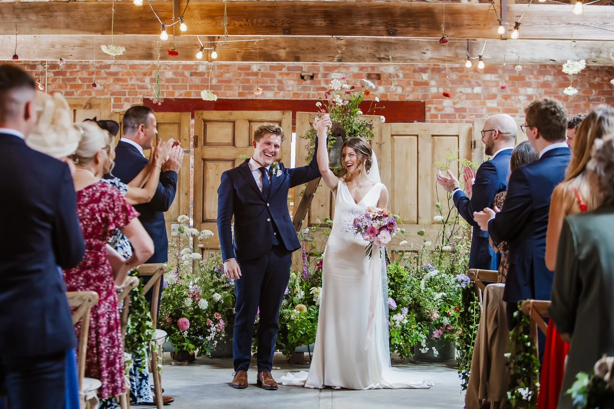 A newly married couple stands hand in hand in front of a decorated backdrop, smiling at each other, surrounded by their guests who are clapping and celebrating. The scene takes place in an indoor rustic wedding venue with wooden beams, brick walls, and hanging lights, with floral arrangements around.