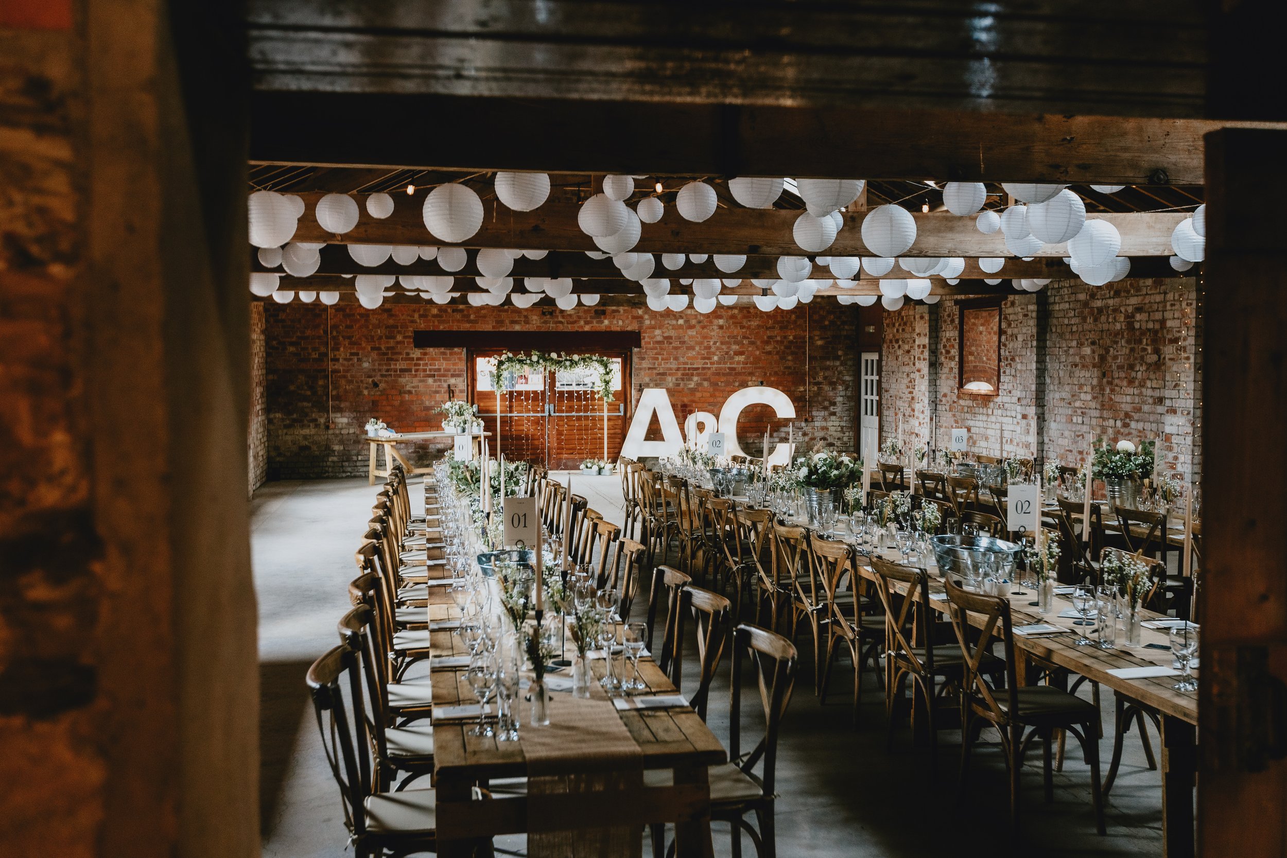 Wedding reception setup with long wooden tables, flowers, and tableware inside a rustic brick venue, decorated with hanging white paper lanterns and large illuminated letters 'A' and 'C' at the back.