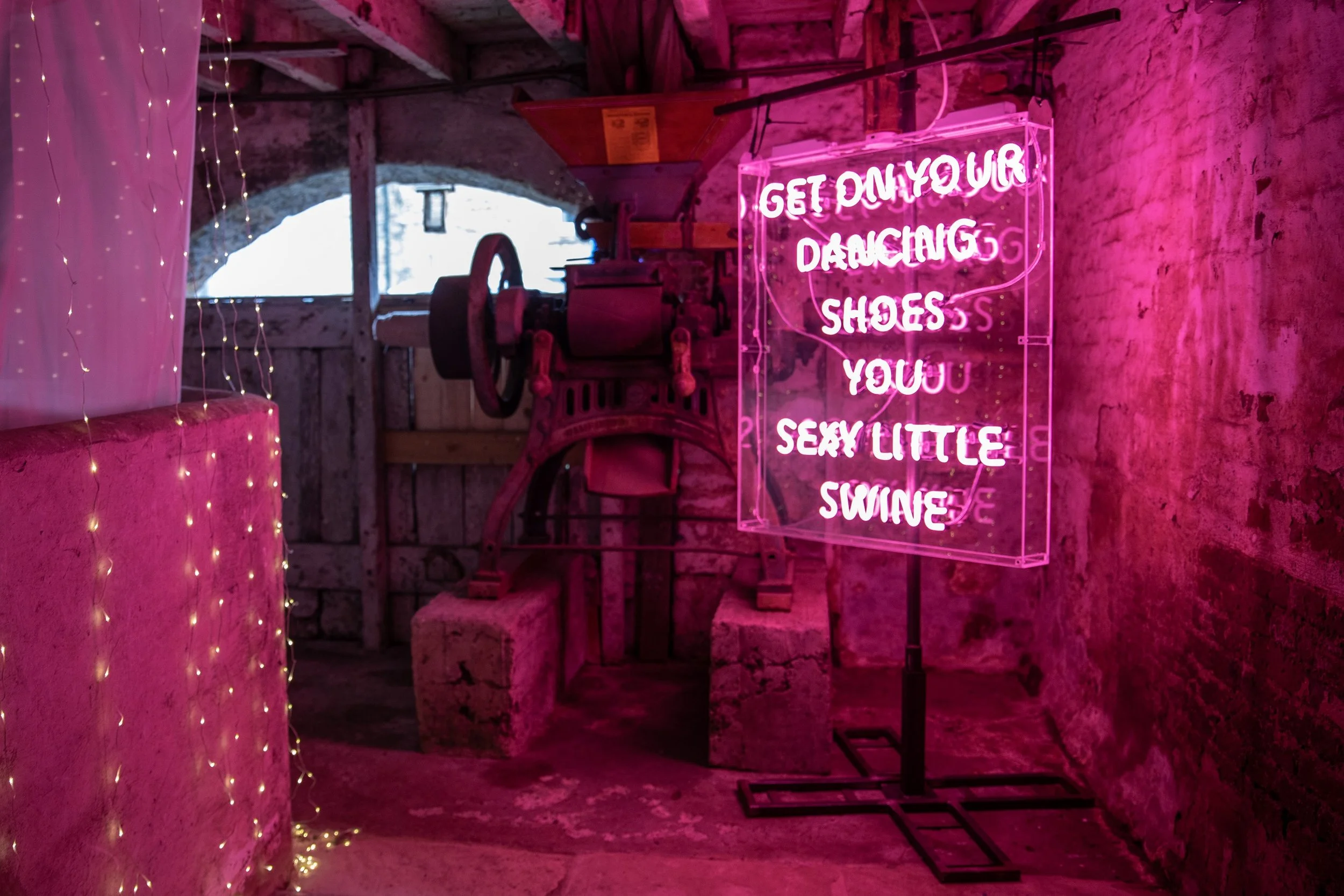 Pink neon sign reading 'Get on your dancing shoes, you sexy little swing' inside a rustic brick room, with string lights and an old machine.