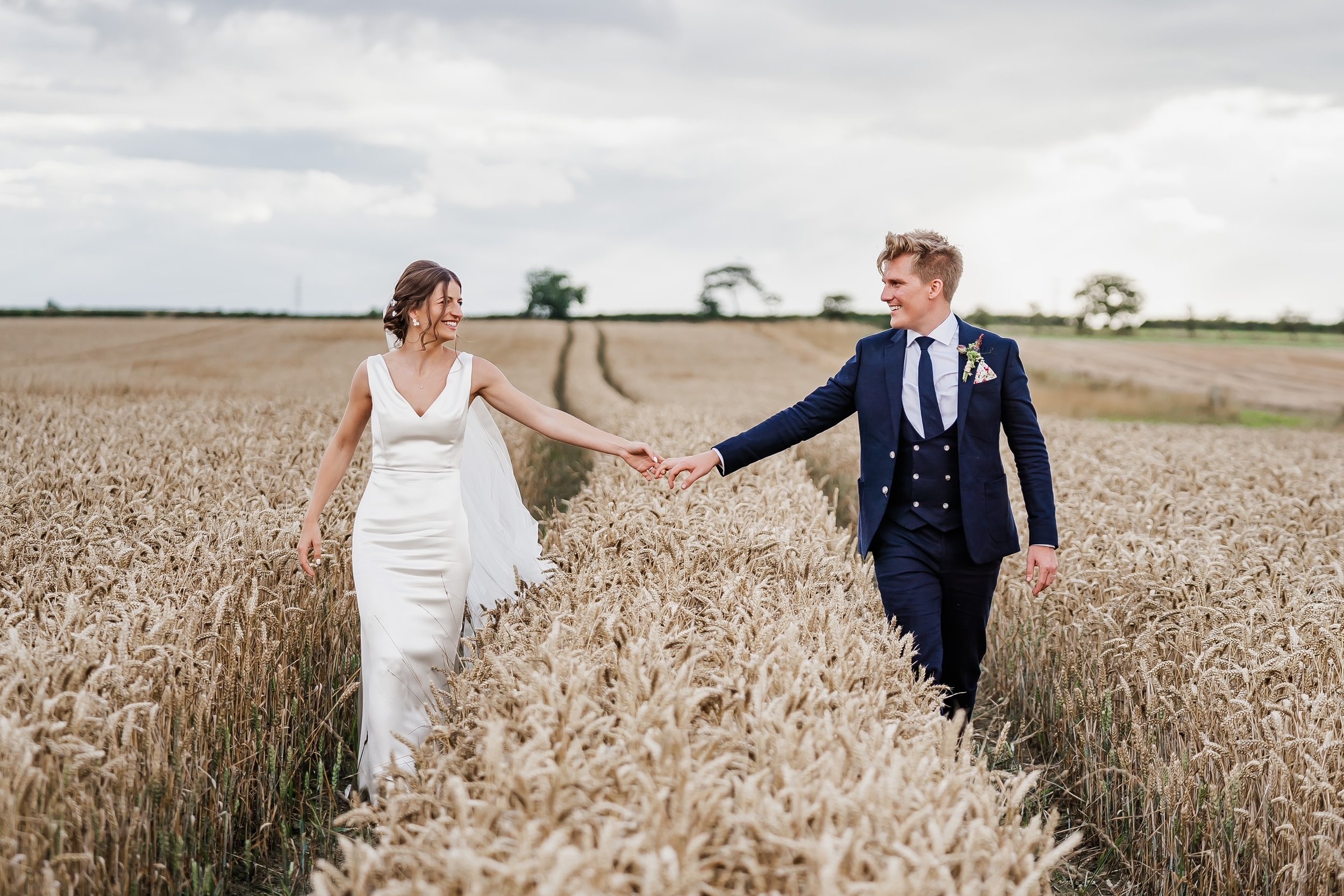 Bride and groom holding hands in a wheat field, smiling at each other, overcast sky.