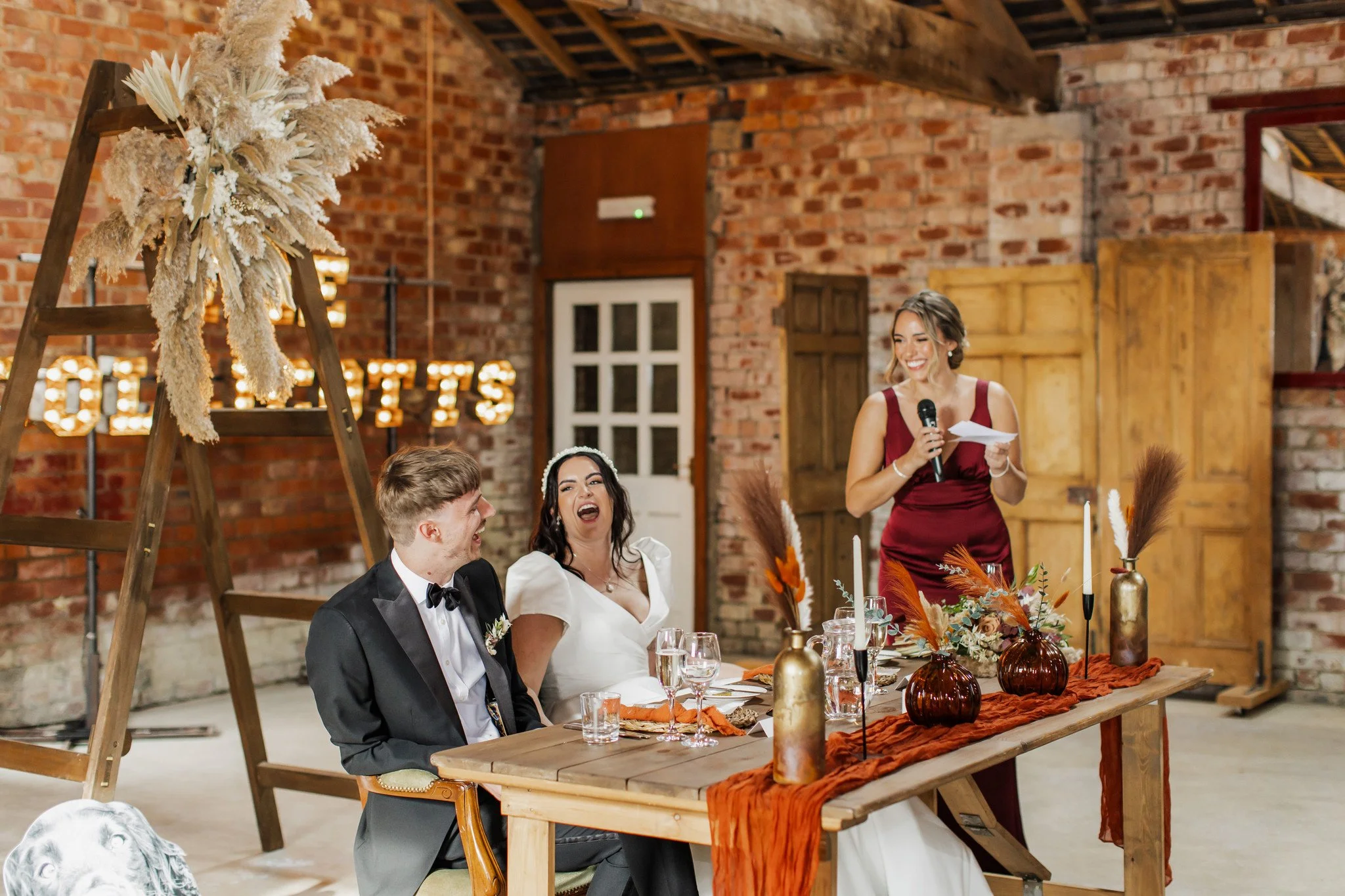 A wedding reception scene with a bride and groom seated at a wooden table, smiling and laughing, while a woman in a red dress gives a speech using a microphone. The table is decorated with orange and brown vases, candles, and floral arrangements, set in a rustic setting with brick walls and a large wooden ladder decorated with dried flowers and pampas grass in the background.