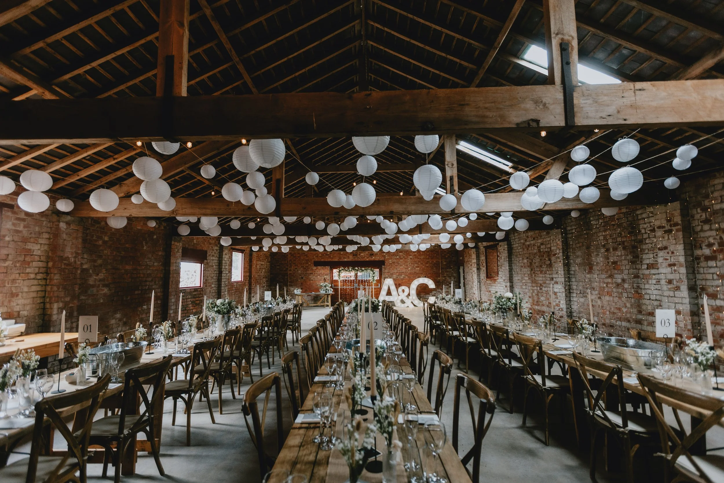 Wedding reception setup in a rustic barn with long wooden tables adorned with white flowers, candles, and glassware. White paper lanterns hang from the wooden ceiling beams, and large white letters 'A & C' are at the far end of the room.
