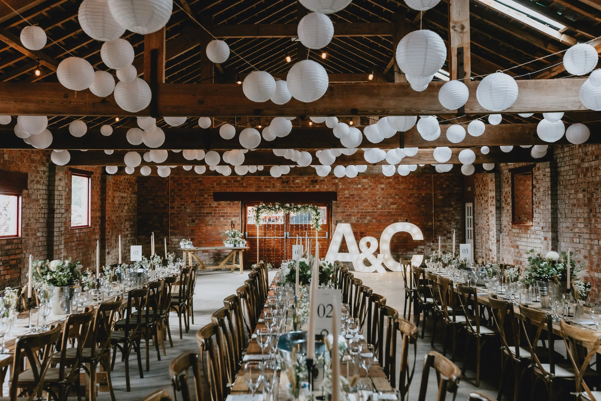 Wedding reception setup in a rustic barn with long dining tables, decorated with floral centerpieces and white candles, white paper lanterns hanging from the ceiling, and large illuminated initials "A & C" at the back of the room.