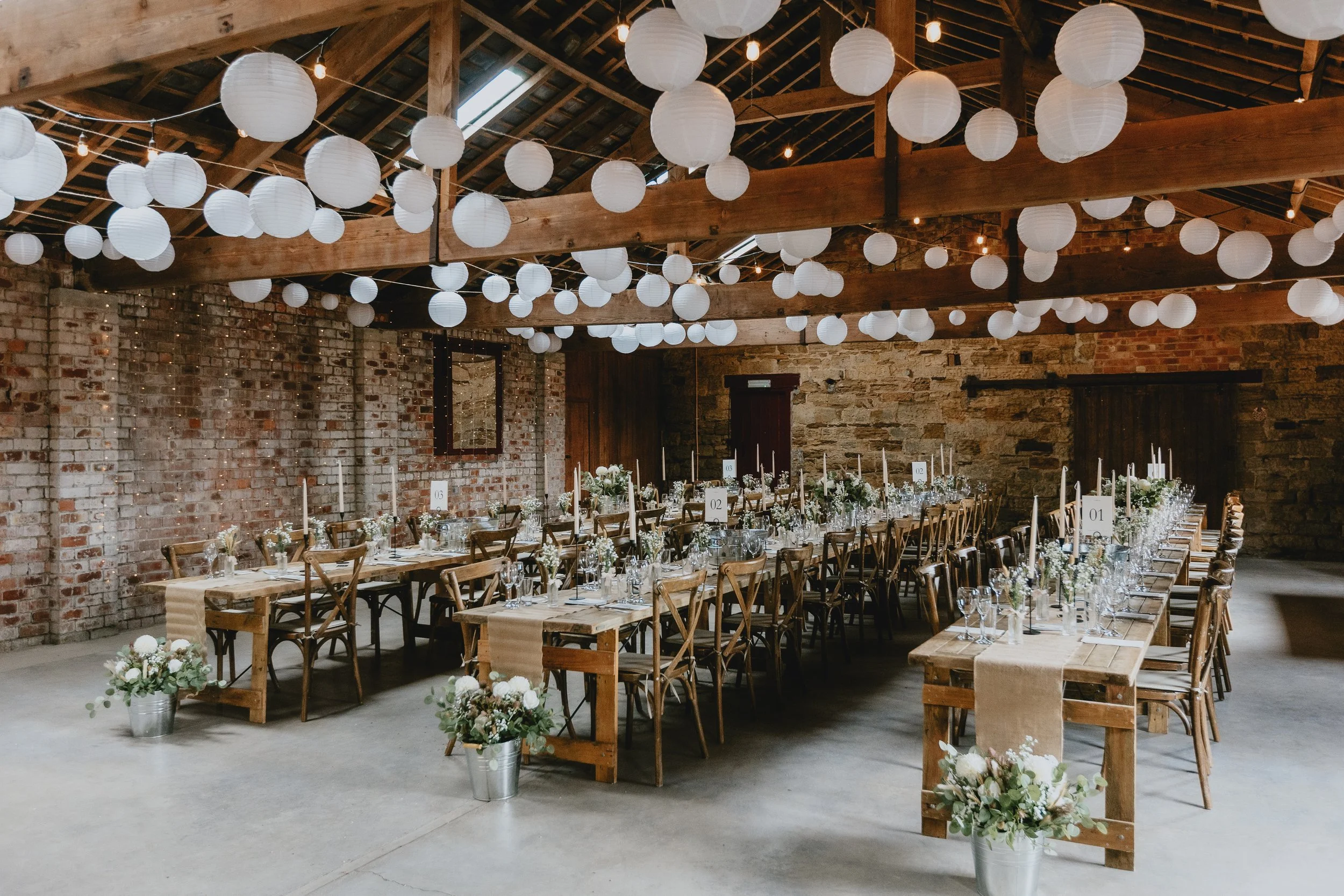 Wedding reception in a rustic barn with wooden tables, chairs, and white flower arrangements, decorated with white paper lanterns hanging from the ceiling.