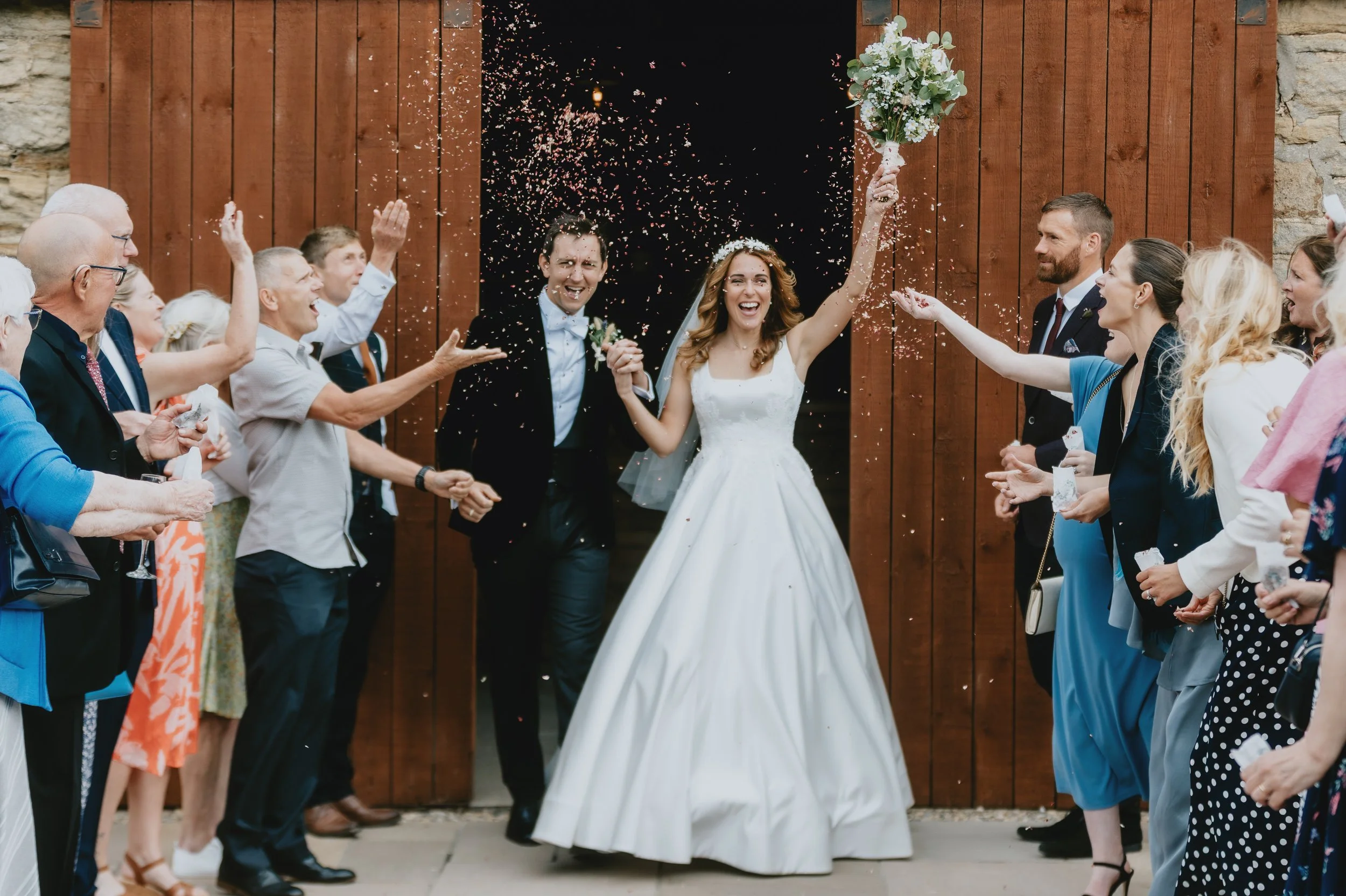 Bride and groom celebrating with guests at their wedding, with confetti in the air and a wooden backdrop.