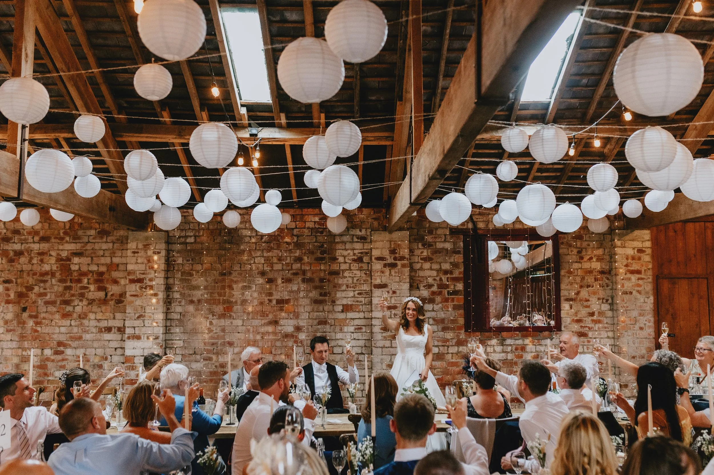 Wedding reception in a rustic barn decorated with white paper lanterns, featuring a bride standing and toasting with guests at a long table.