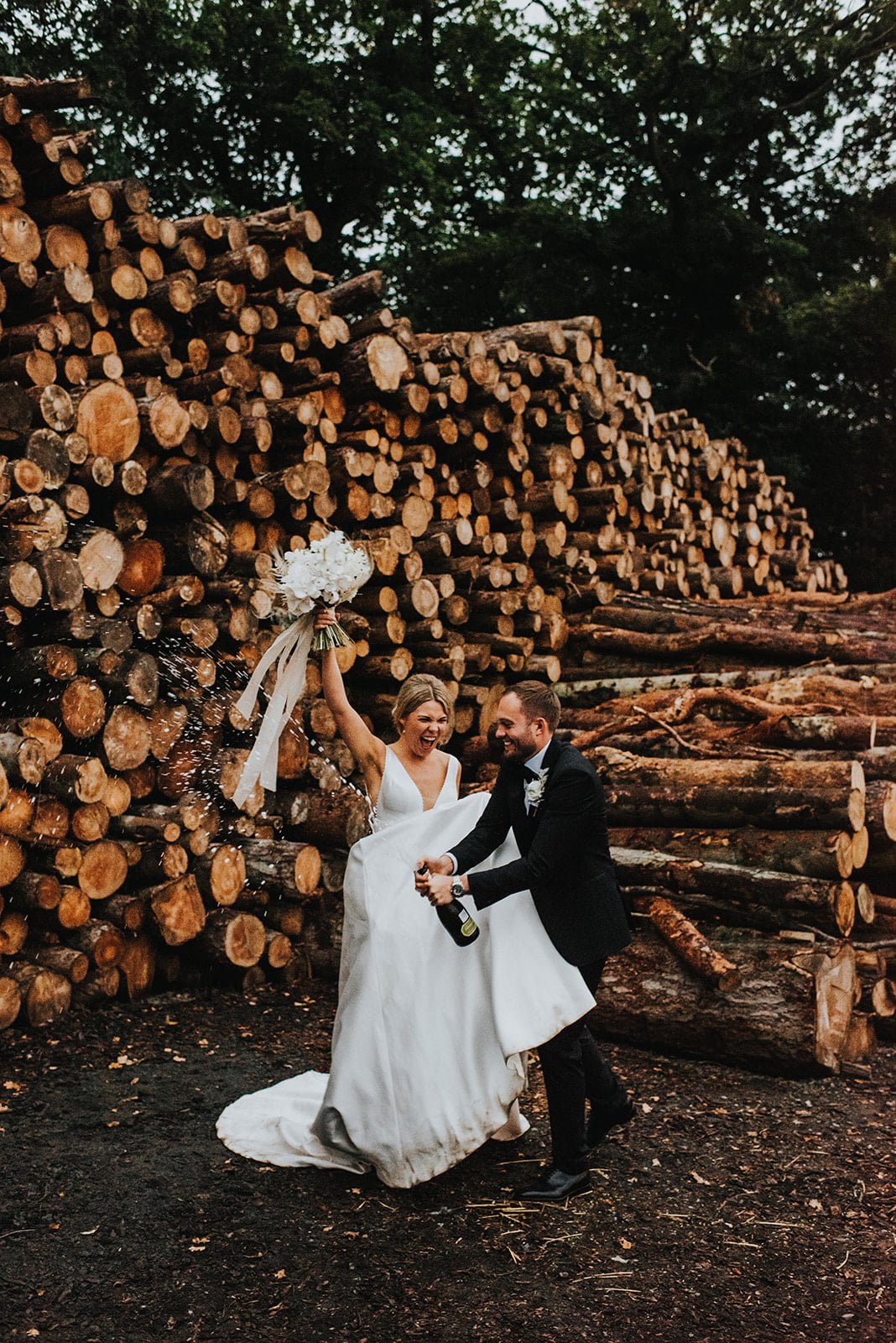 Bride and groom celebrating outdoors with a large stack of chopped firewood in the background. The bride is holding a bouquet and appears joyful, while the groom is holding a drink from a bottle.