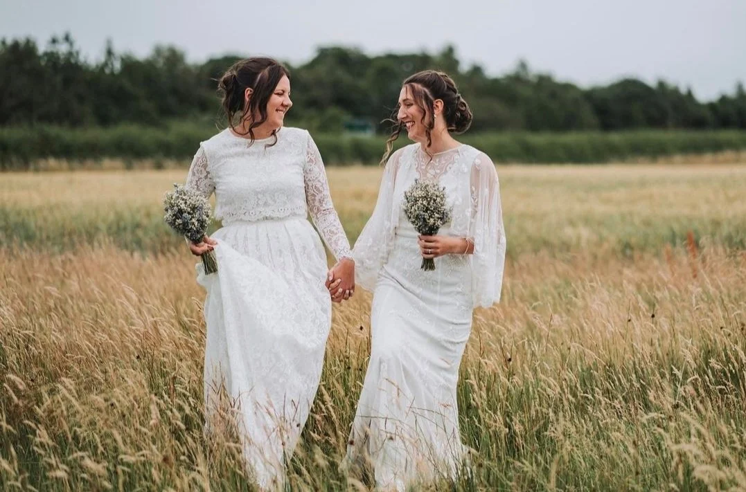 Two women in white wedding dresses holding hands and smiling in a field of tall grass, each holding a bouquet of white flowers.