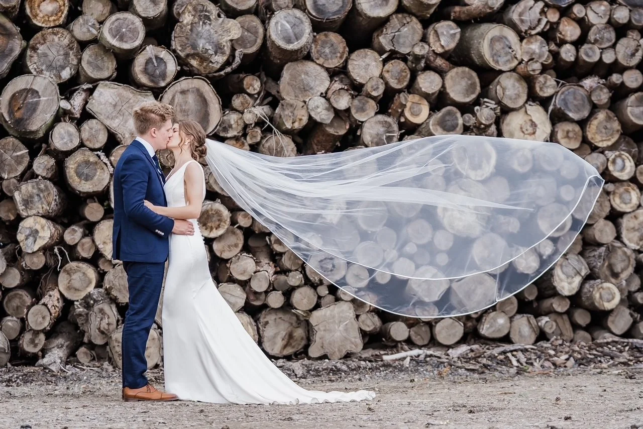 A bride and groom kissing outdoors, with a large stack of chopped logs in the background. The bride is wearing a white wedding gown and veil, and the groom is dressed in a blue suit.