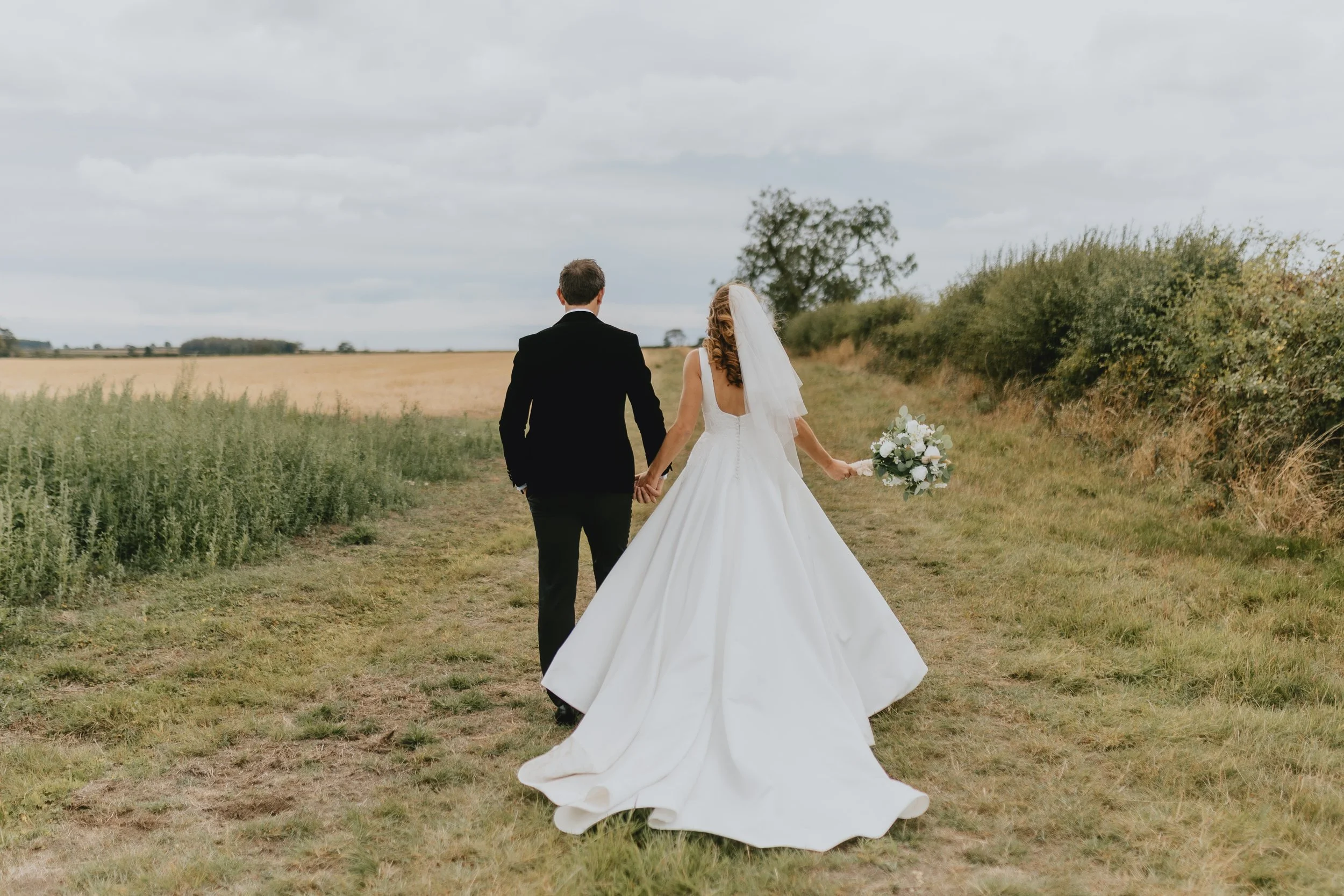 Bride and groom walking hand in hand through a field, with the bride holding a bouquet, seen from behind on a cloudy day.