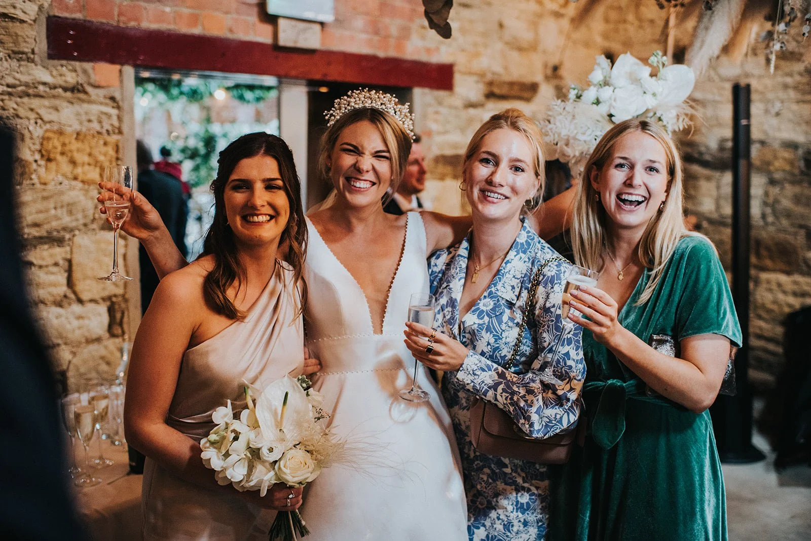 Group of four women celebrating at a wedding reception, smiling, holding drinks, with one wearing a bridal veil and crown, in a rustic indoor setting.