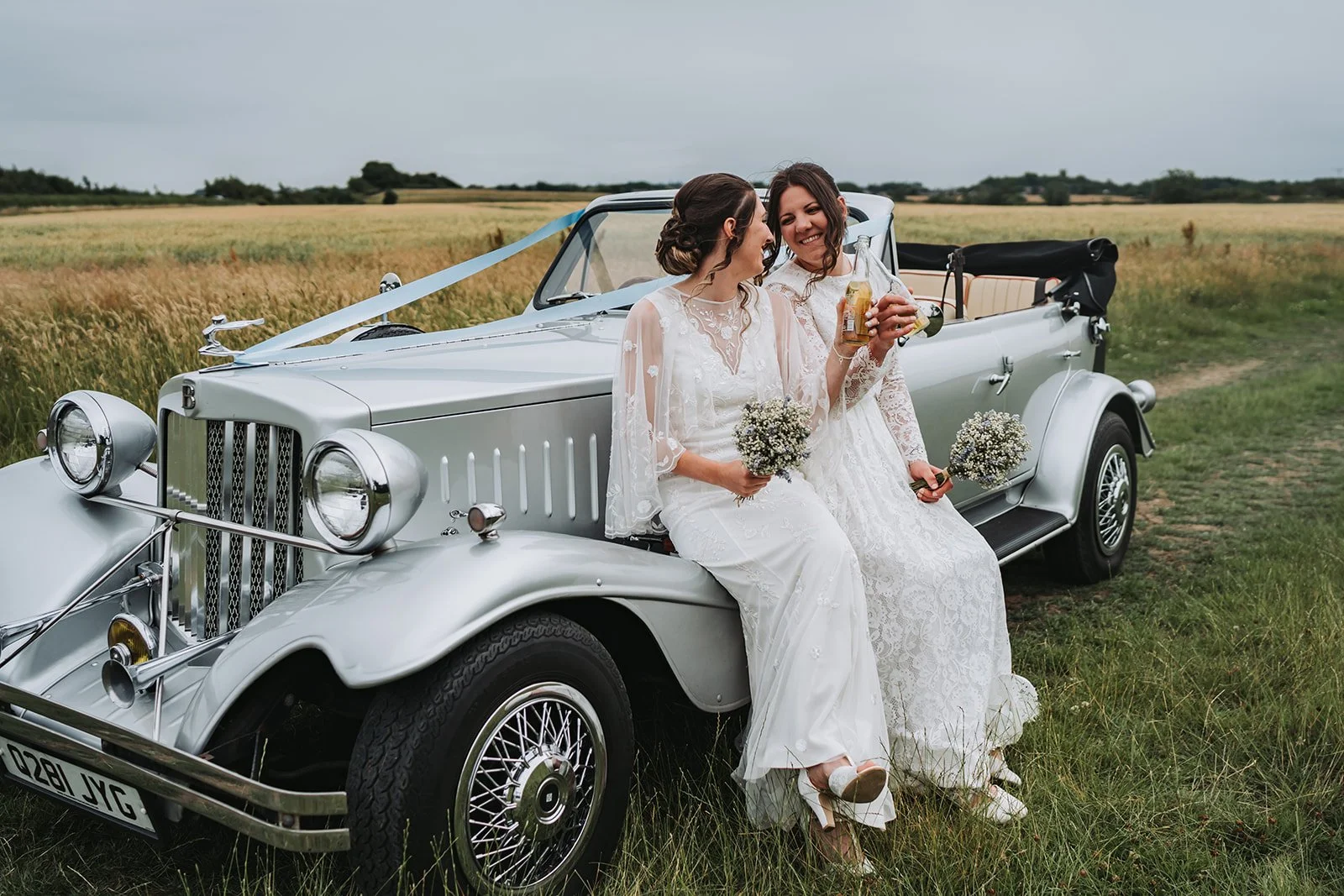 Two women in wedding dresses sitting on the front of a vintage white convertible car in a field, holding bouquets and drinks, smiling at each other.
