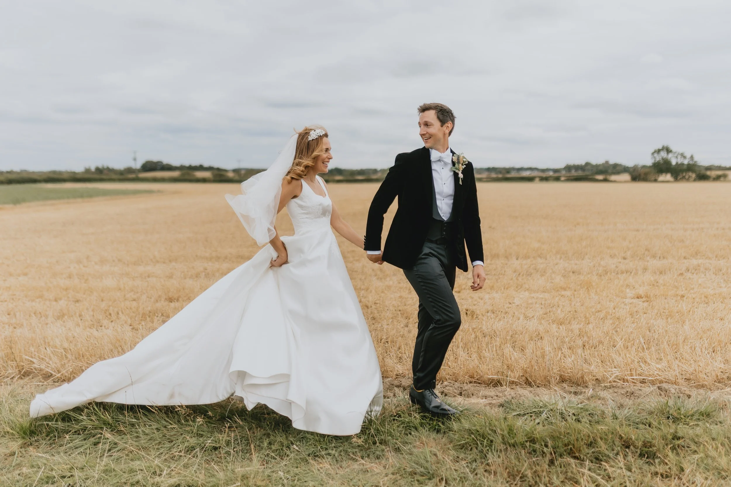 Bride and groom in wedding attire, holding hands and smiling, walking across a grassy field with a golden crop in the background and overcast sky.