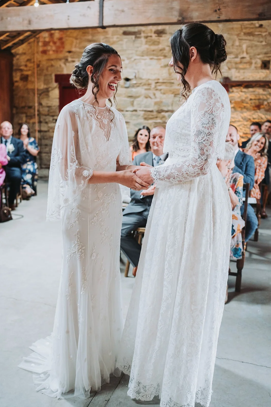 Two brides holding hands and exchanging vows during a wedding ceremony, with guests seated and watching in a rustic venue.