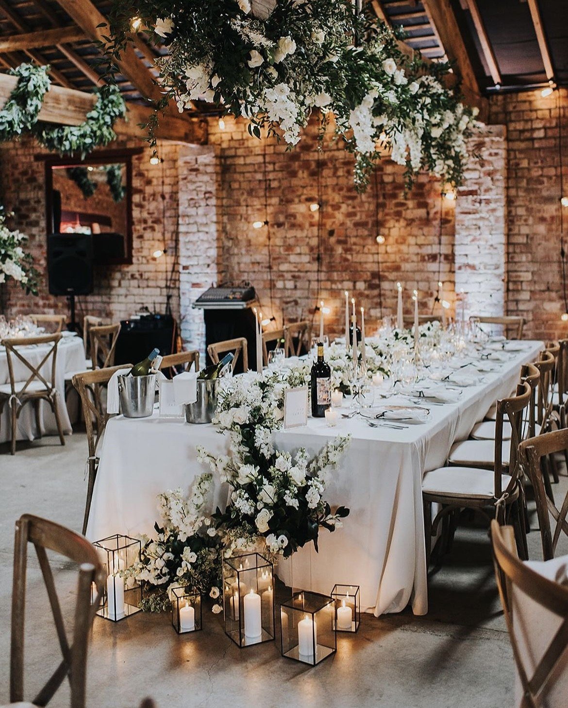 Elegant wedding reception setup with a long white dining table decorated with white floral arrangements, tall white candles in candelabras, and wine bottles. The venue features exposed brick walls, wooden beams, string lights, and hanging greenery.