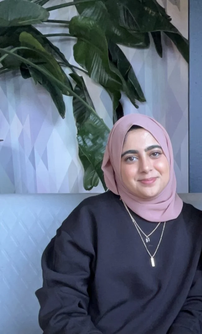 A woman wearing a light pink hijab and black top, sitting on a light blue cushion, with green tropical leaves in the background.