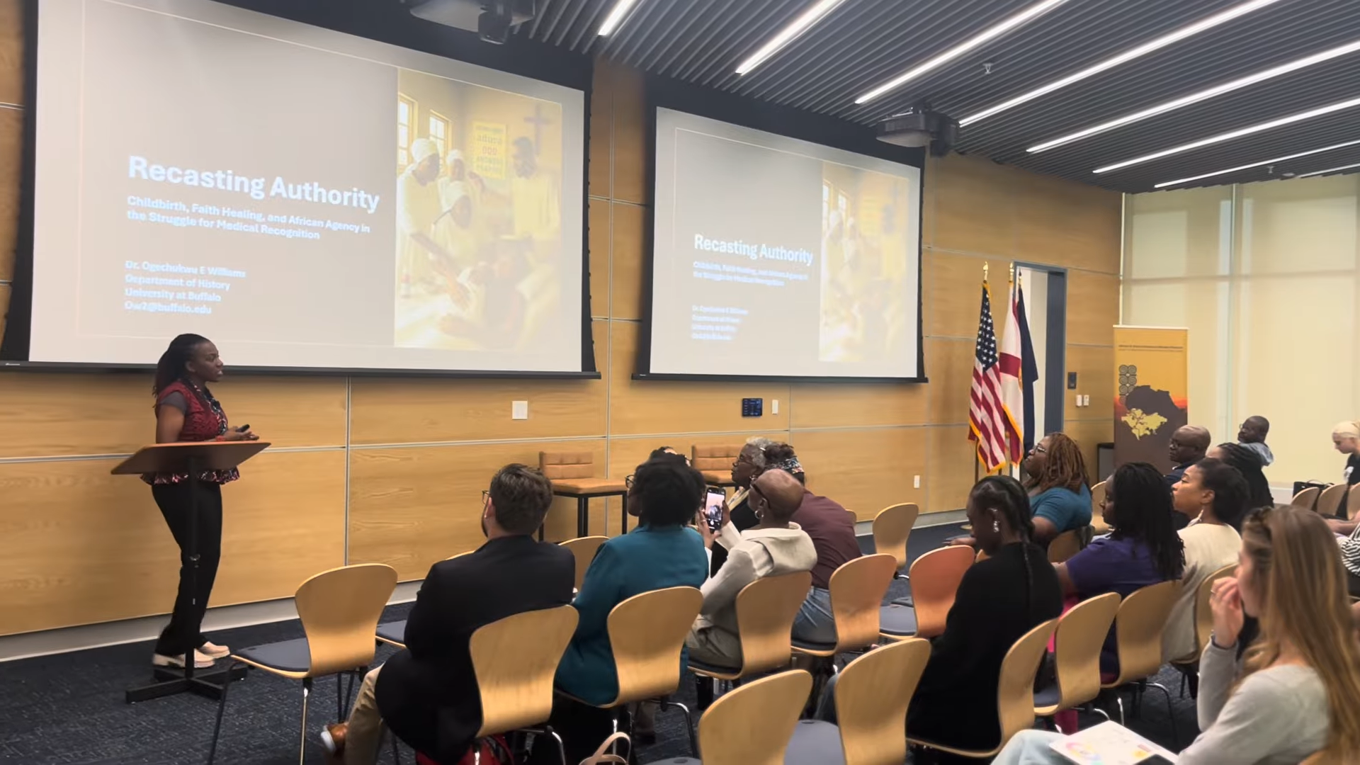 A woman presenting at a conference in a room with an audience. The presentation slide behind her reads 'Recasting Authority: Childbirth, Faith Healing, and African Agency in the Struggle for Medical Recognition' and shows an image of people worshiping. The room has wooden walls, two projection screens, and flags of the United States and another country.