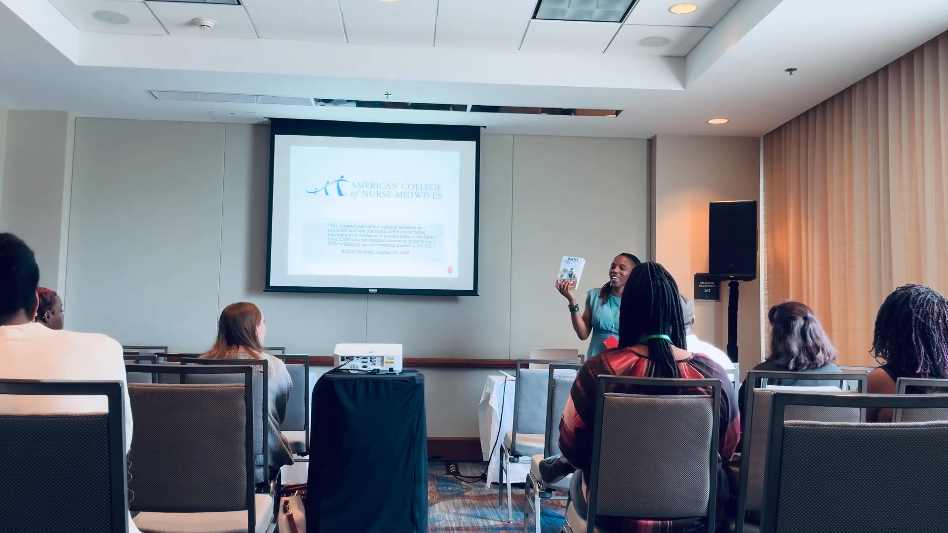 A woman is giving a presentation to a seated audience in a conference room. The presentation slide on the screen displays the logo of the American College of Nurse-Midwives.