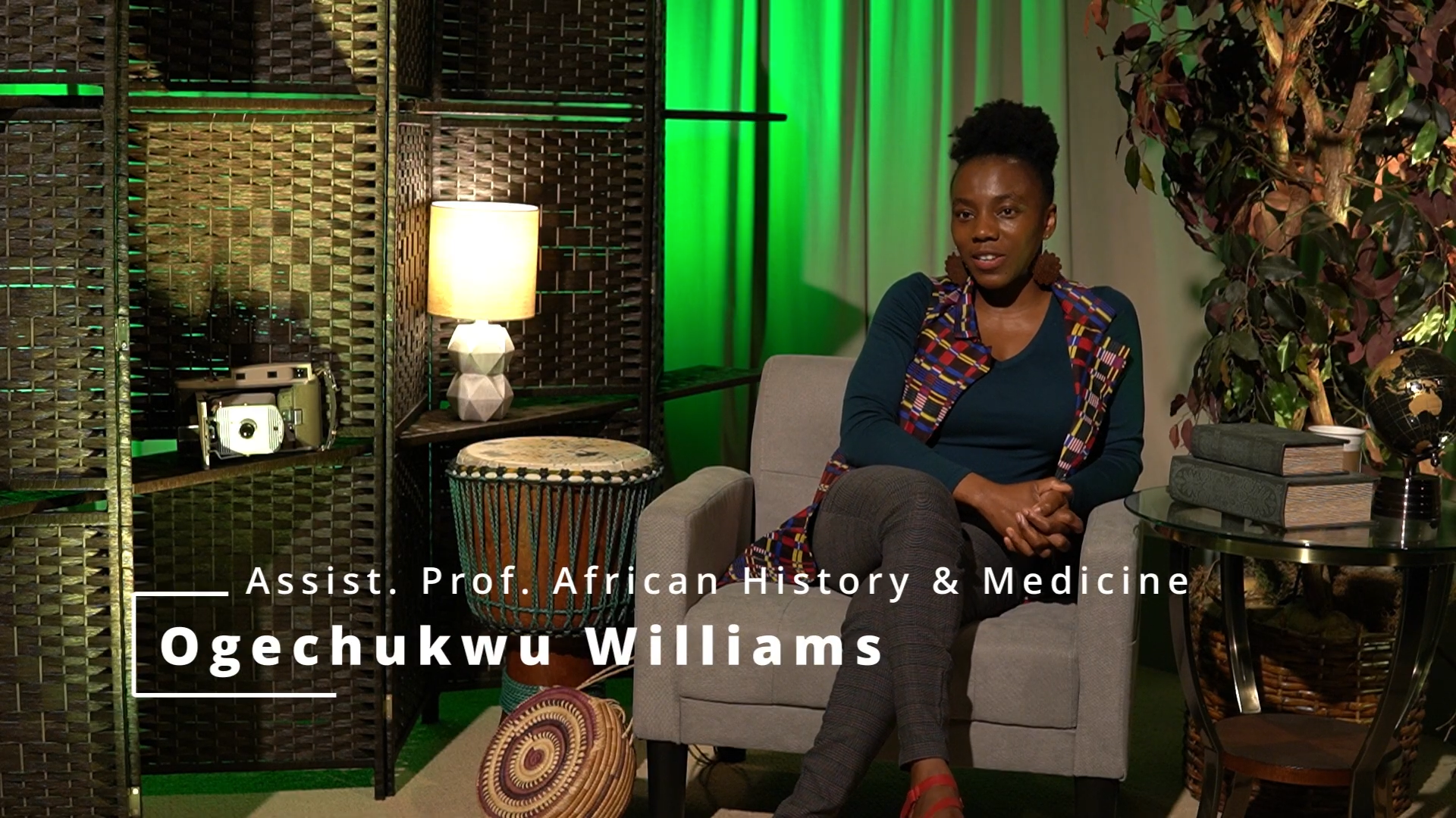 An African woman sits on a beige armchair in a studio with decorative shelves, green lighting, books, plants, and a globe visible in the background.