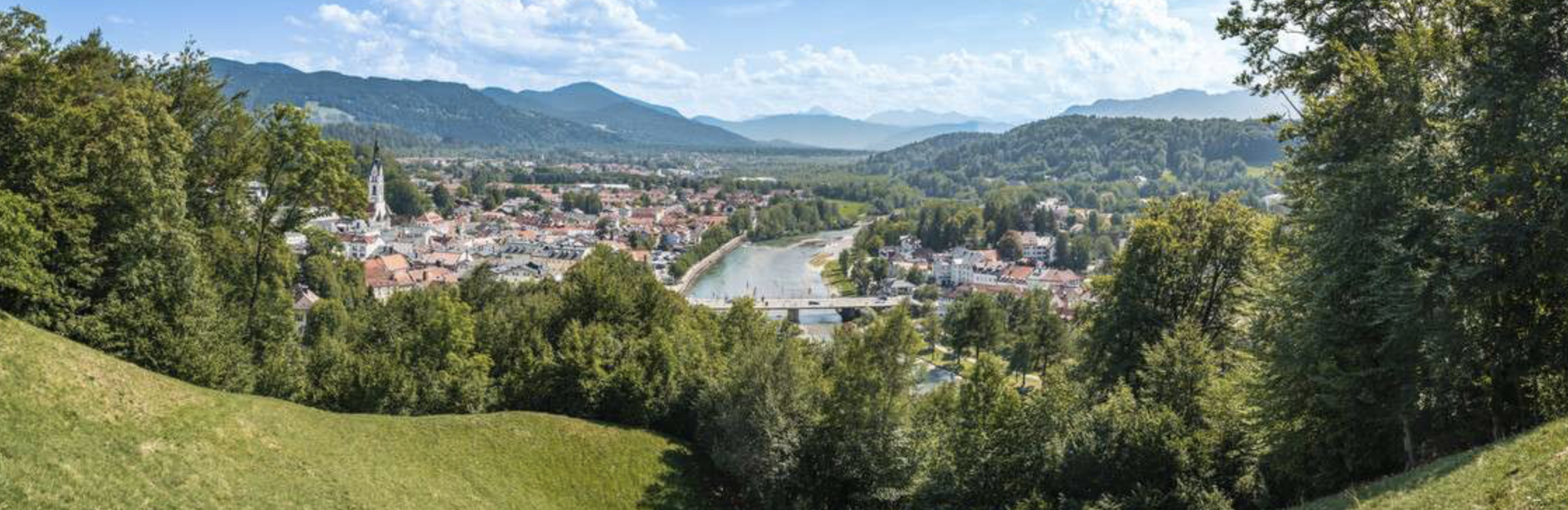 Panoramablick auf eine Stadt in einer flachen Flussebene, umgeben von grünen Hügeln und Bergen im Hintergrund, mit einem Fluss, der durch die Stadt fließt, und Brücken. Viele Häuser und eine Kirche sind sichtbar.