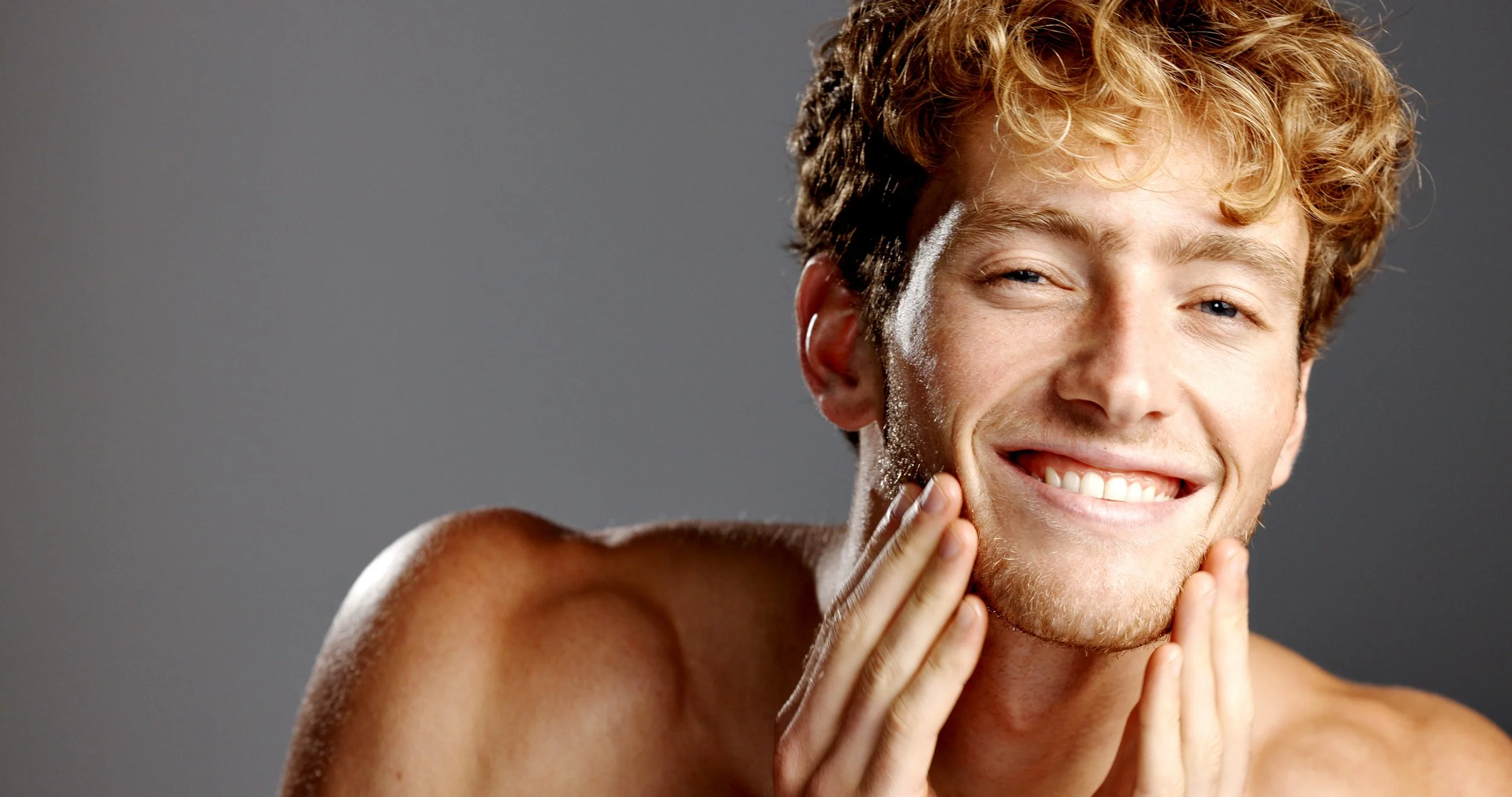 Ein junger Mann mit lockigem, hellbraunem Haar, der sich im Gesicht rasiert und lächelt.