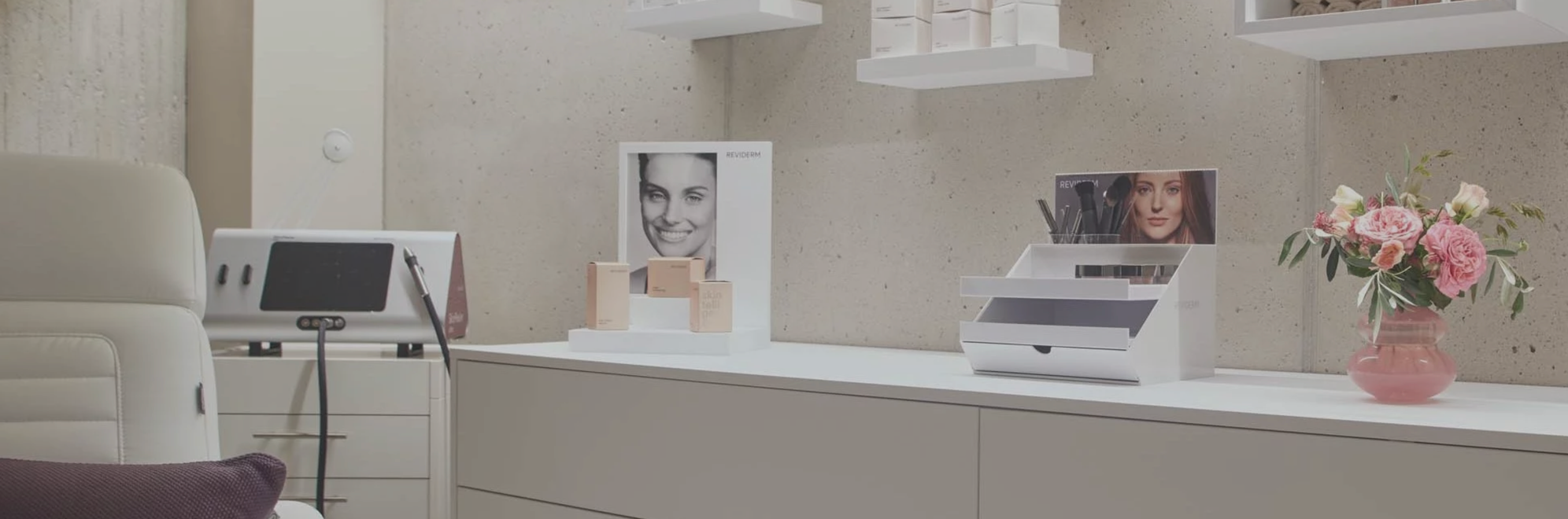Ein weißer Behandlungsraum mit einer weißen Liege und Kosmetikprodukten auf einem weißen Schreibtisch, einem Bilderrahmen mit einem lächelnden Gesicht, Make-up-Pinsel in einem weißen Organizer, und einer Vase mit pinken Blumen auf der rechten Seite.