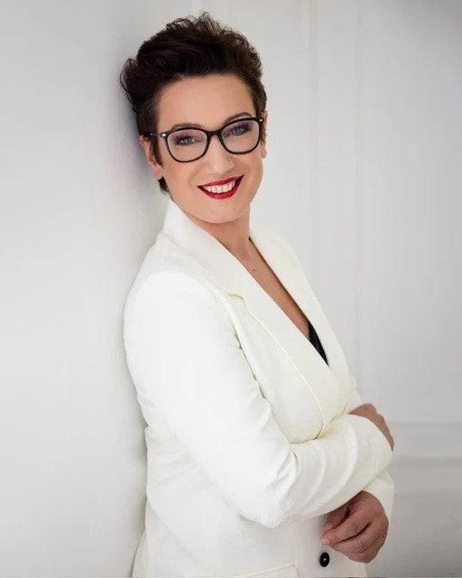 Eine Frau mit kurzen dunklen Haaren, Brille und rotem Lippenstift, die in einem weißen Blazer lächelnd steht, vor weißer Wand.