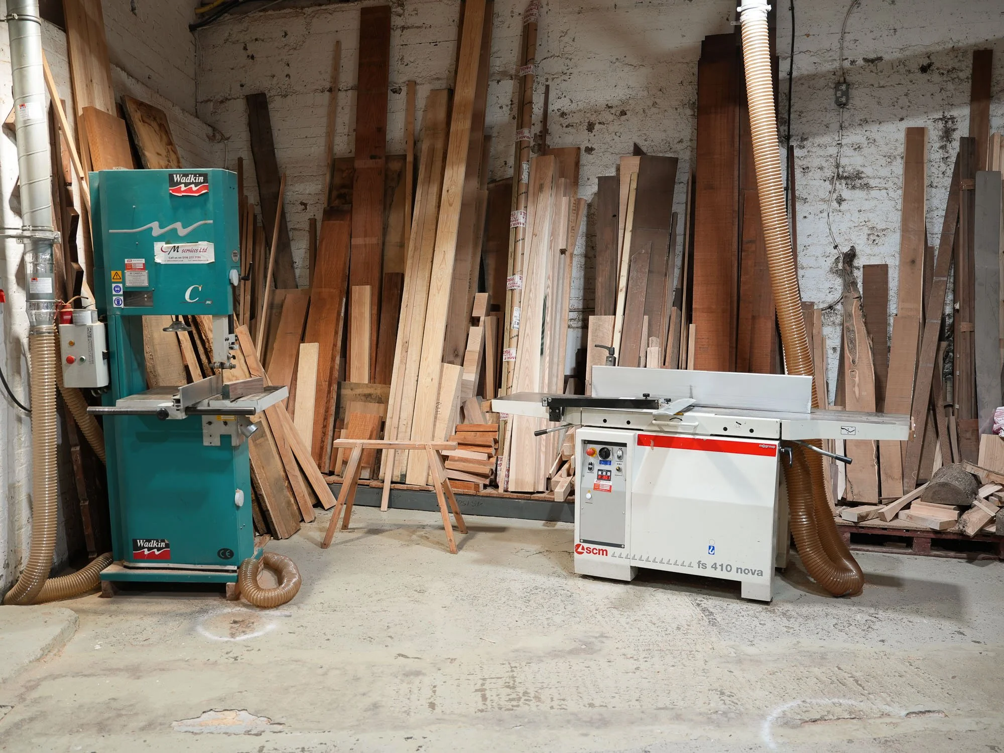 A woodworking workshop with various pieces of wood leaning against the wall, a saw machine, and a dust extraction system.
