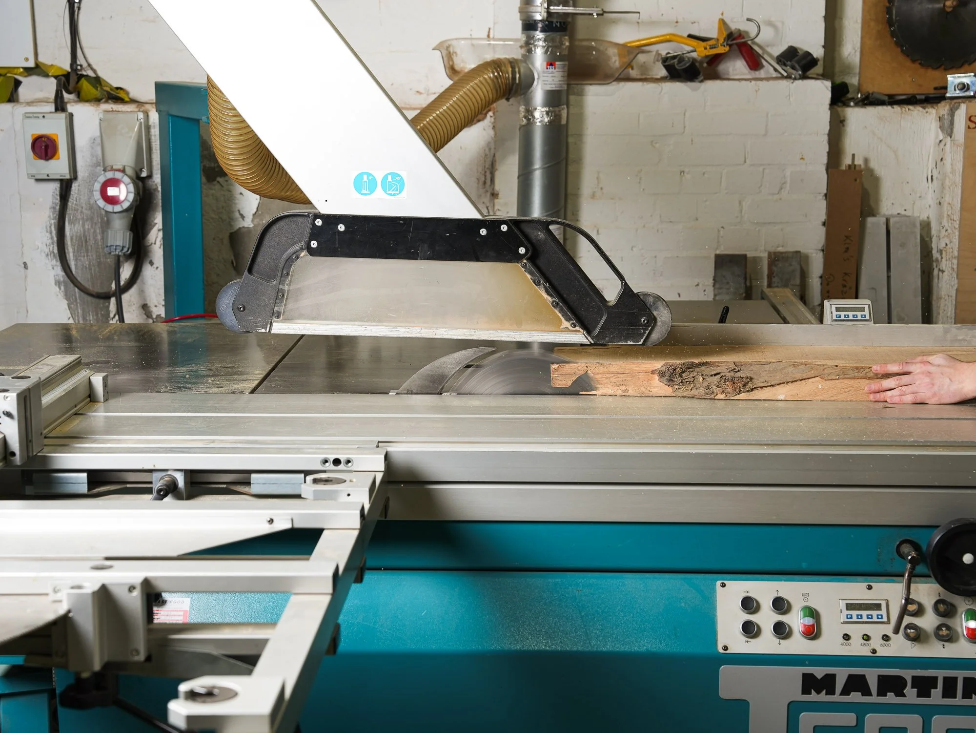 A woodworking workshop showing a blue and gray table saw cutting a piece of wood, with various tools and equipment in the background.