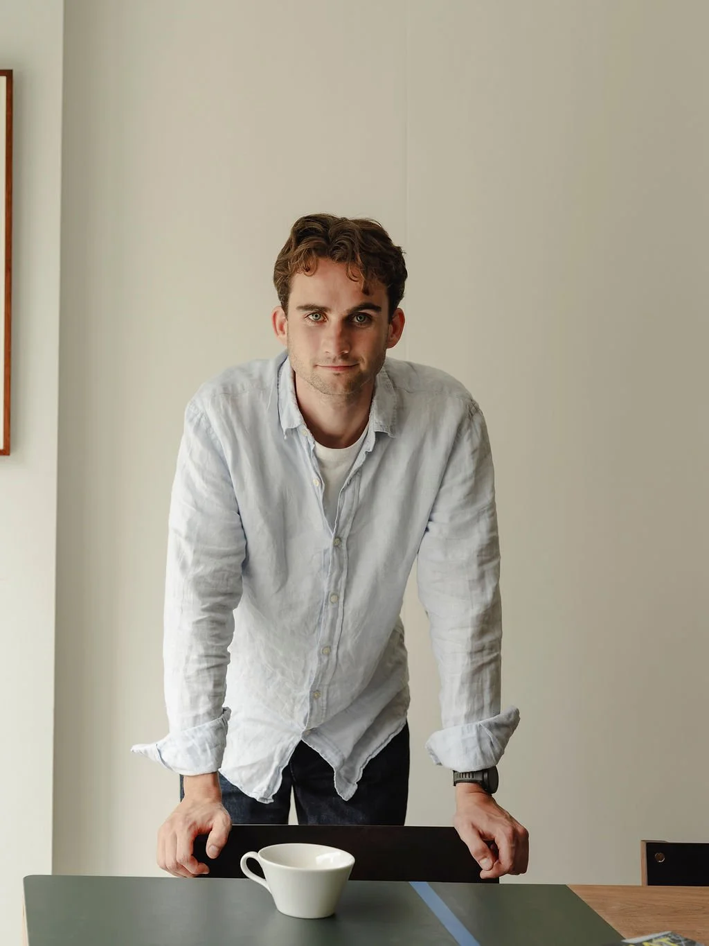 A young man leaning on a table with a white coffee cup, wearing a light-colored shirt, in a minimalistic room with cream walls.