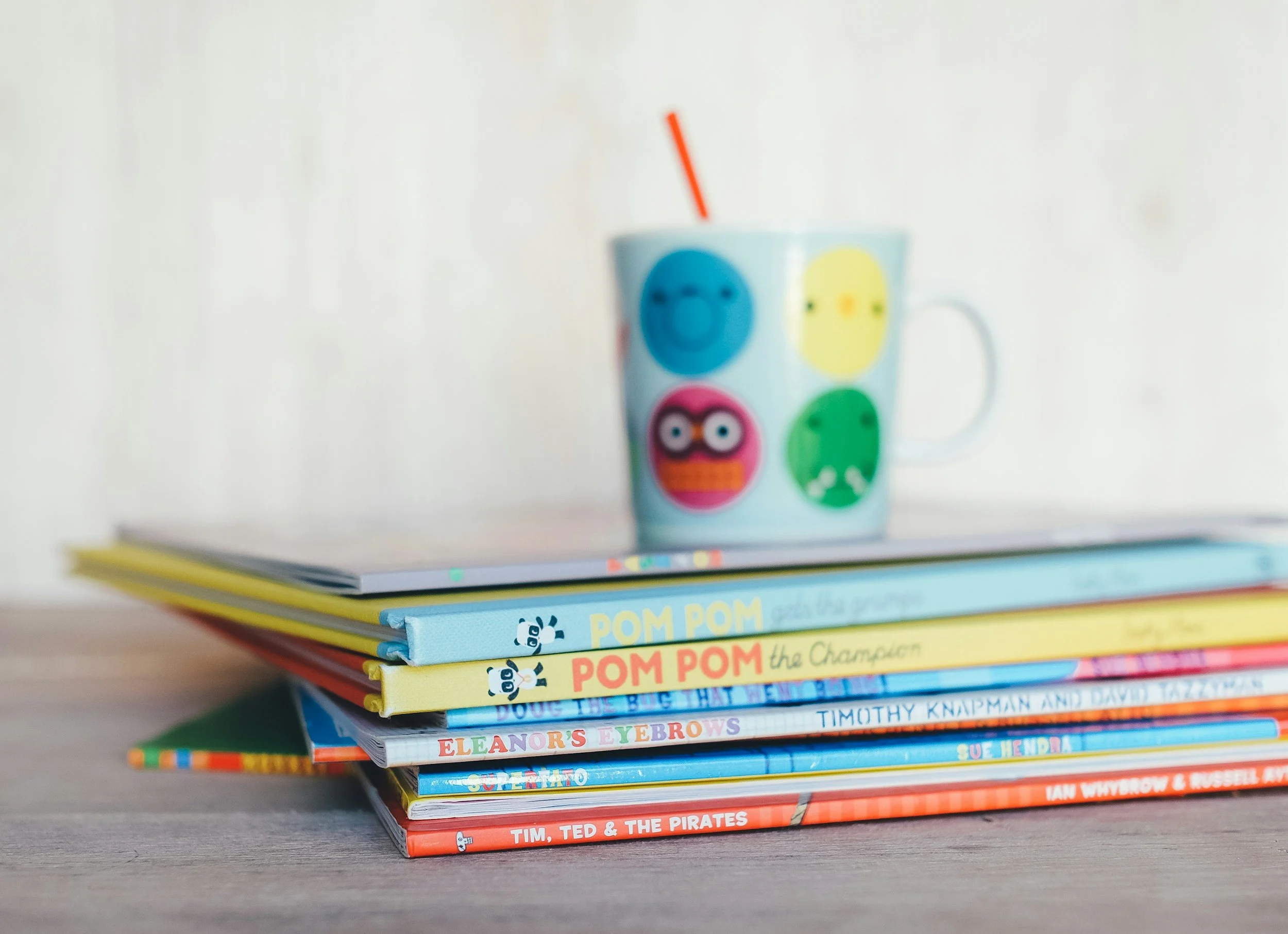 Stack of colorful children's books on a wooden surface with a white mug decorated with cartoon animal faces in the background.