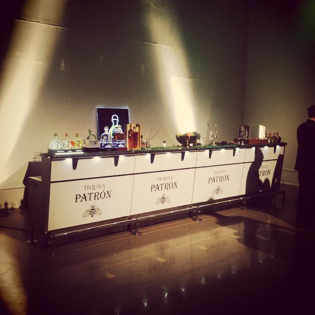 Long bar table covered with a white cloth that has 'TEQUILA PATRÓN' printed on it, with liquor bottles and bar accessories on top, in a dimly lit room.
