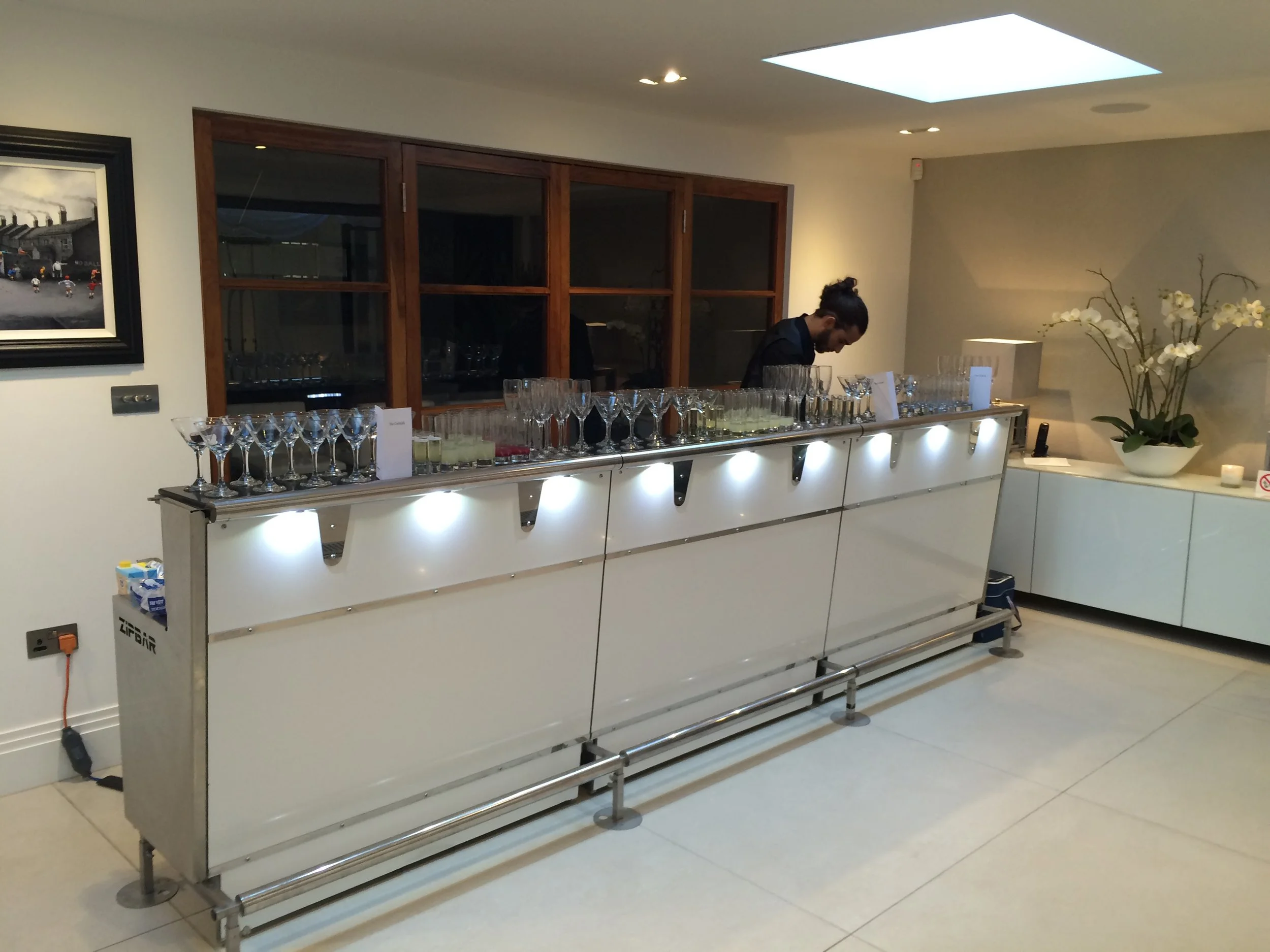 A bartender behind a white bar with glasses and drinks, preparing beverages in a modern indoor setting with artwork, large window, and orchids.