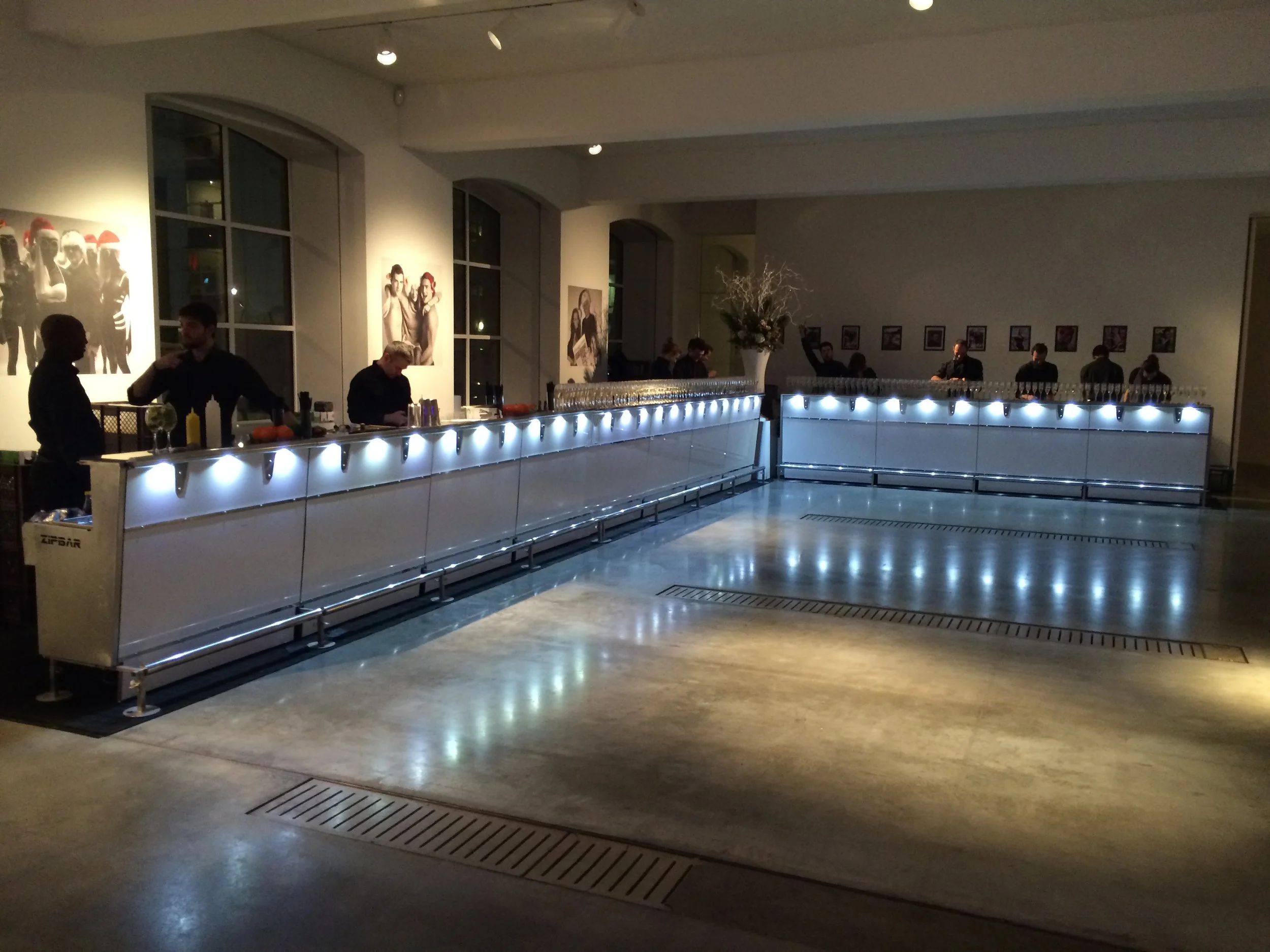 Empty bar with illuminated counter at a party or event venue, with some people setting up or working behind the bar and framed photos or artwork on the walls.
