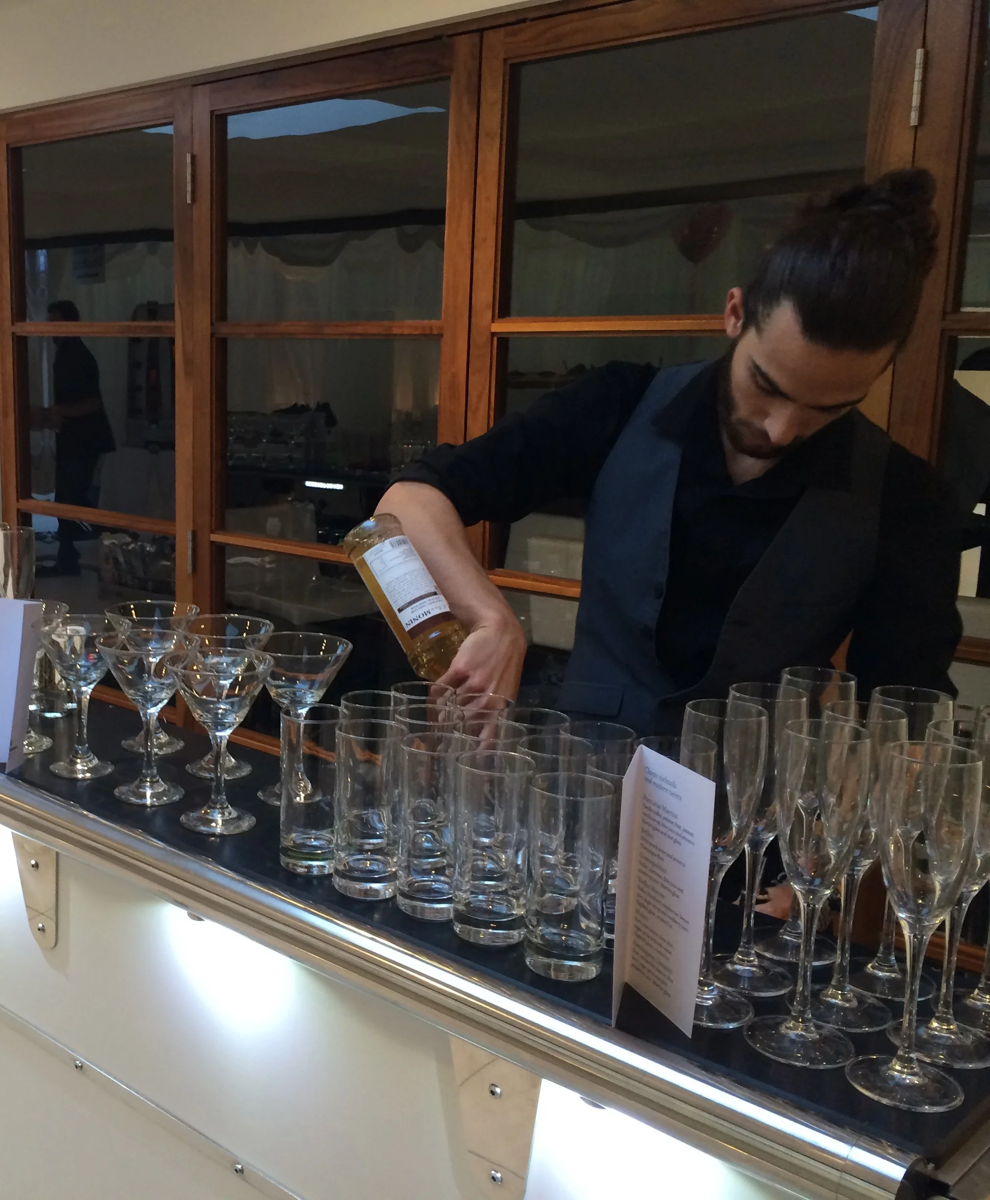 A bartender pours a drink into a row of empty cocktail glasses at a bar or event setup.