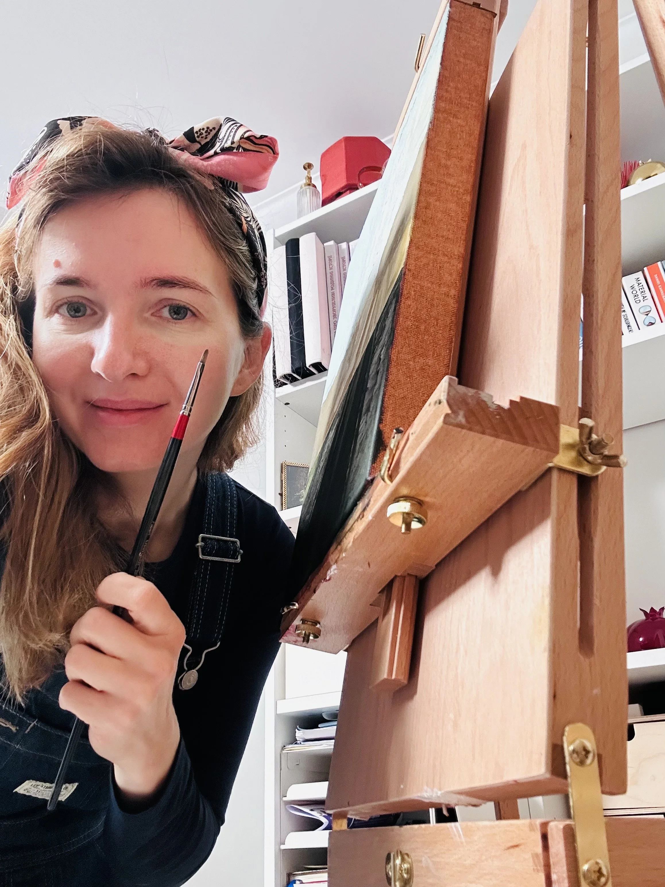 A woman with light brown hair, wearing a colorful headscarf and a black shirt, is holding a paintbrush next to a small wooden easel with a canvas, in a room with shelves filled with books and decorative items.