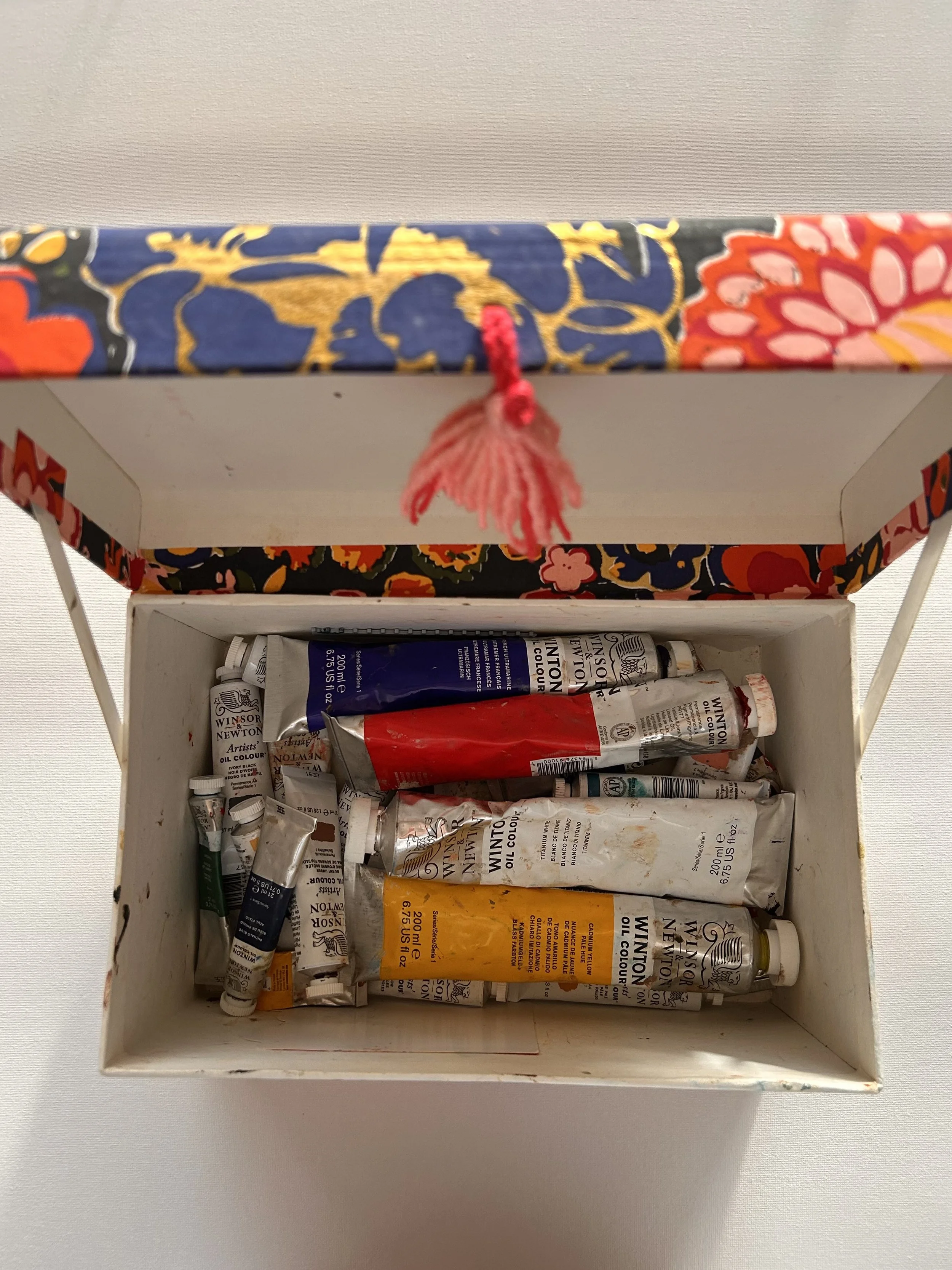 Open box containing used oil paint tubes in various colors, with a decorated fabric lid.