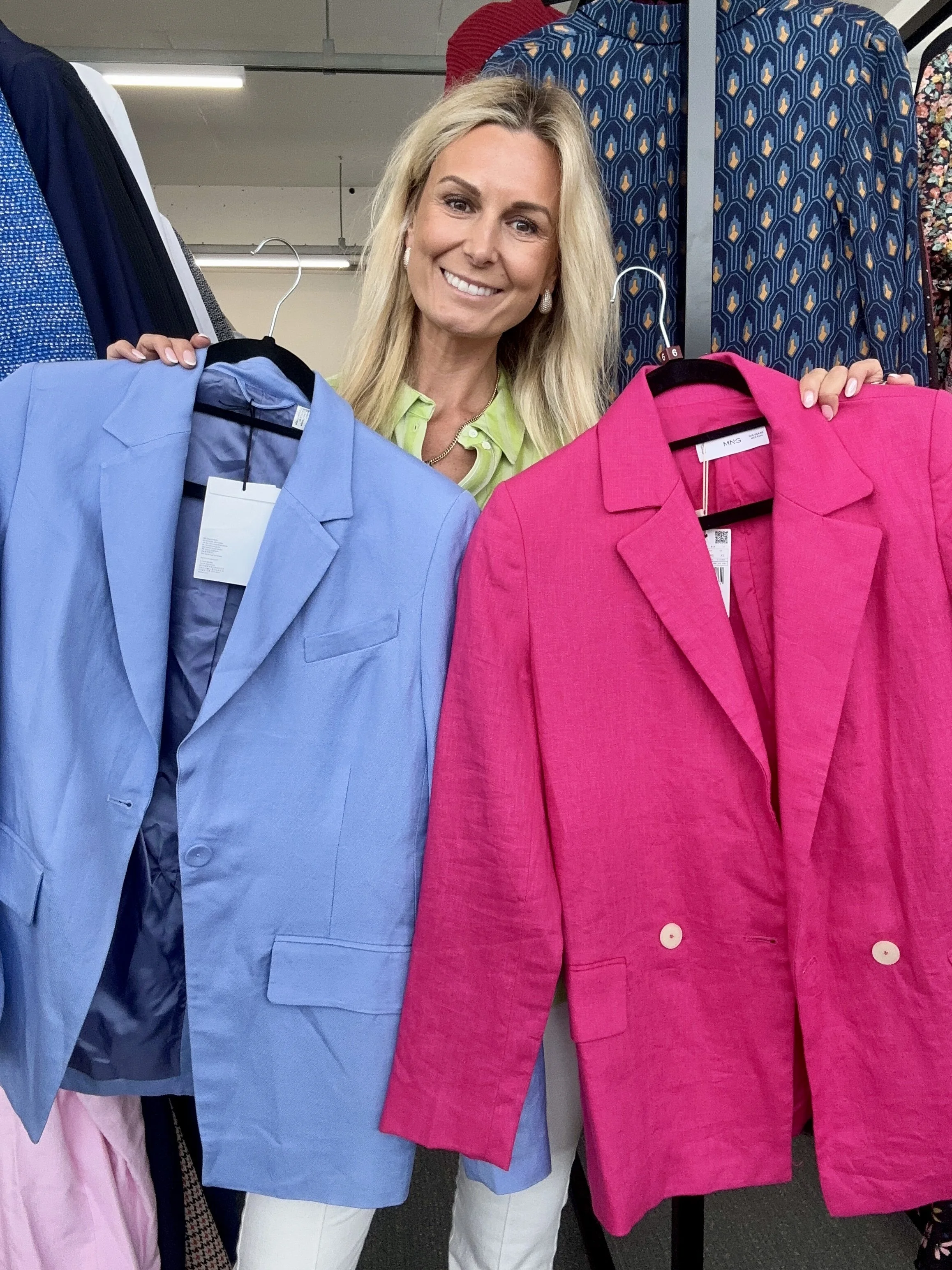 A woman with blonde hair smiling while holding two blazers, one light blue and one bright pink, in a clothing store.