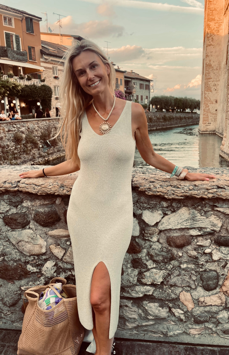 A woman with long blonde hair in a cream-colored dress with a slit, standing by a stone railing near a canal with colorful buildings and a cloudy sky in the background.