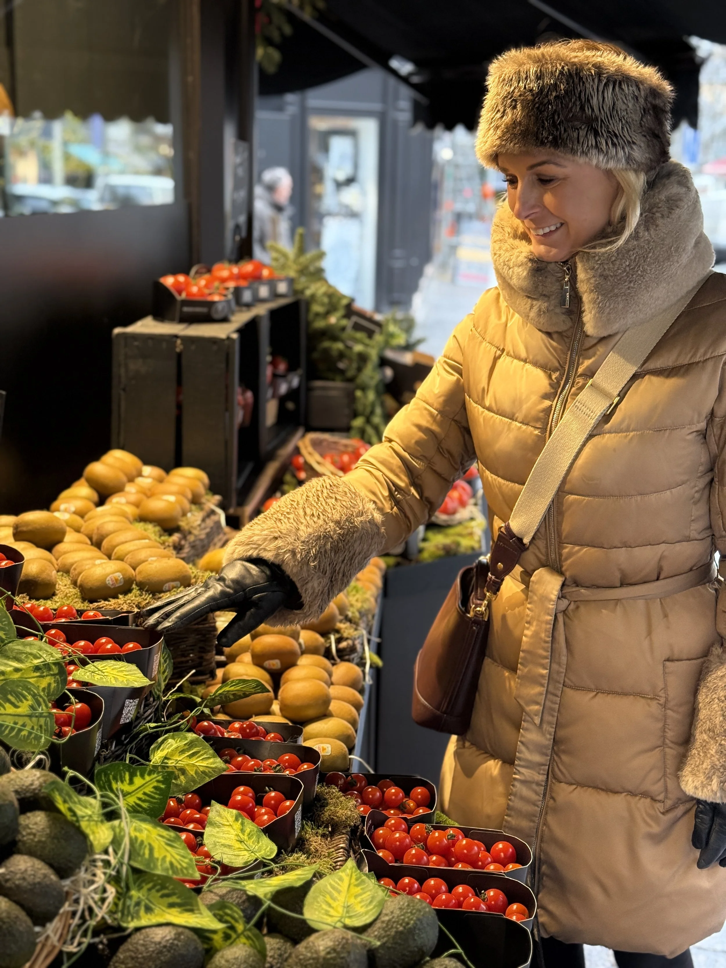 A woman shopping for fresh tomatoes and other vegetables at an indoor farmers market. She is wearing a beige coat with a fur-lined hood, and black gloves, and is smiling as she looks at the produce.