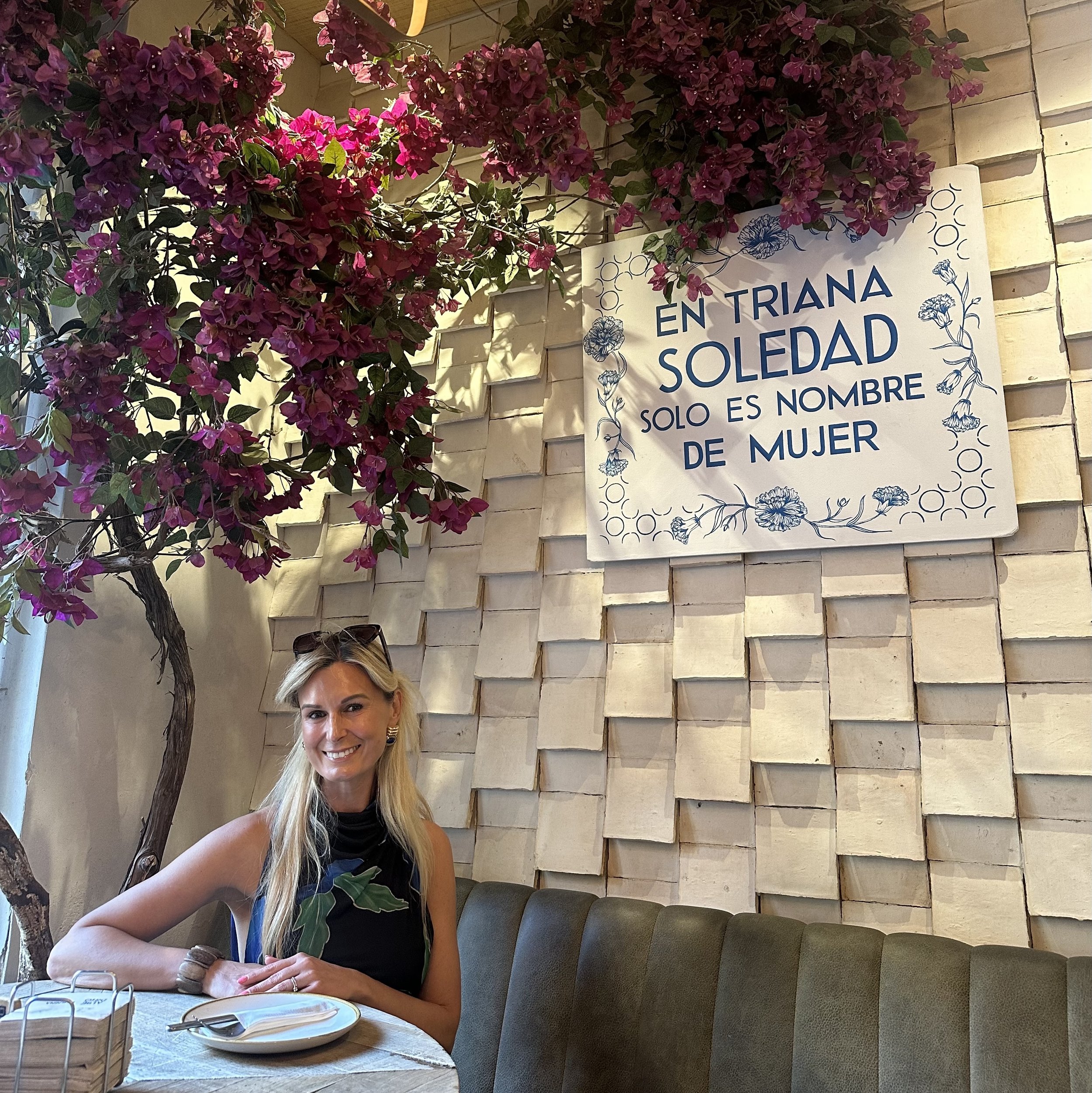 A woman with blonde hair smiling, sitting at a dining table in a restaurant, with a background of a textured wall and pink bougainvillea flowers.