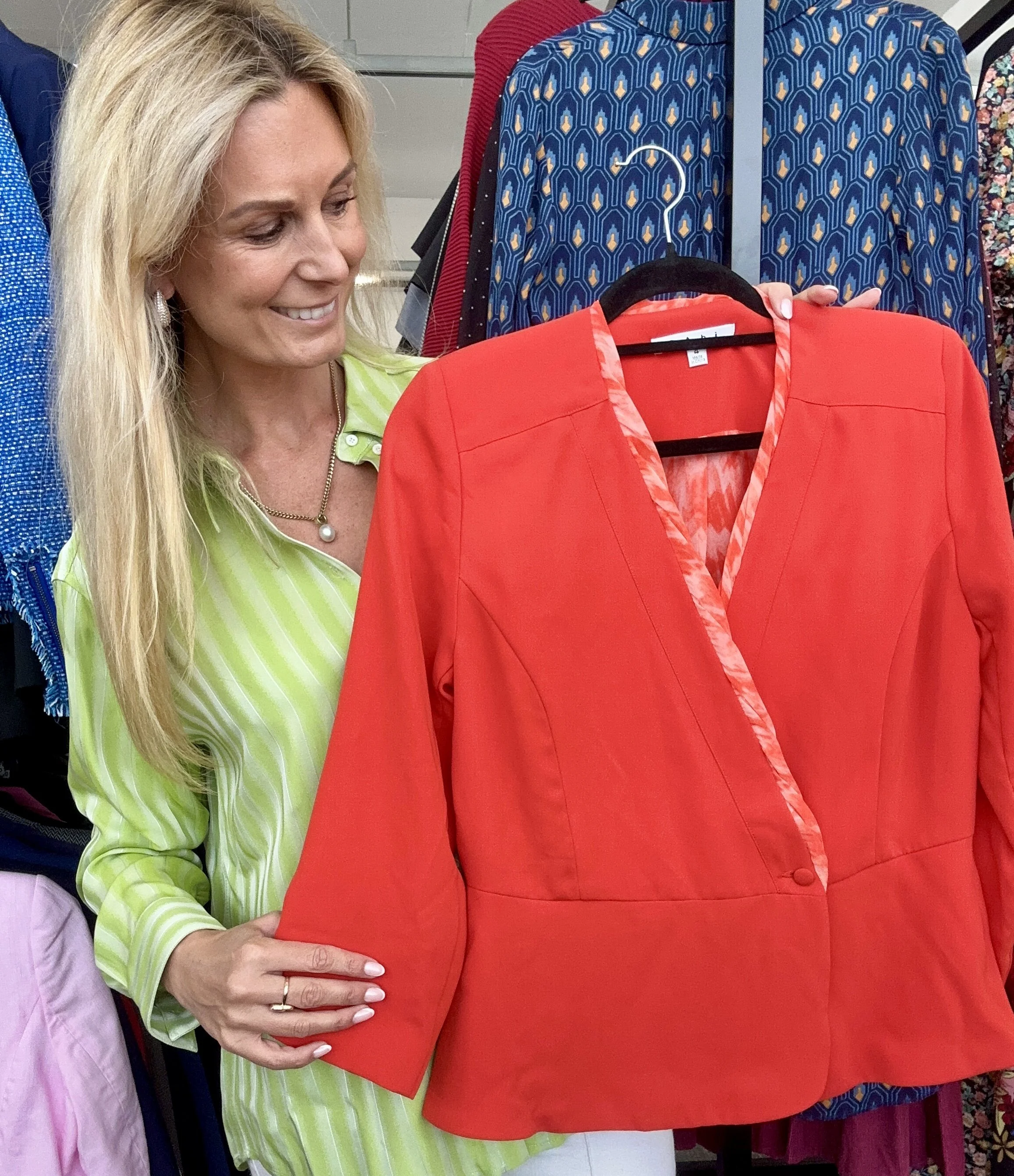 A woman with blonde hair smiling while looking at a red blazer on a hanger. She is holding the blazer with one hand and wearing a green and white striped shirt with a pearl necklace.