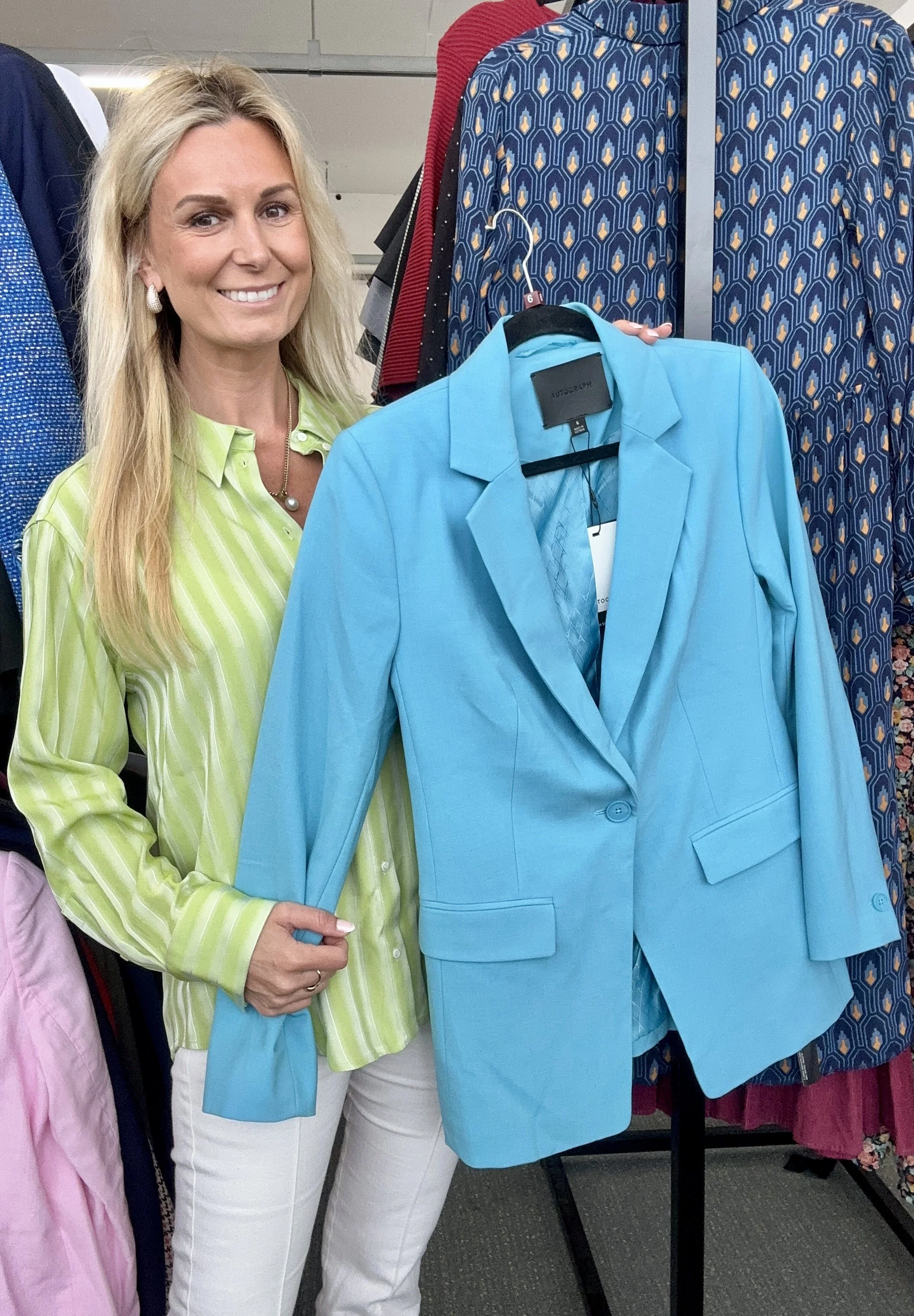 Vivienne with long blonde hair smiling at the camera, holding a cyan blazer on a hanger in a clothing store. She is wearing a light green and white striped shirt and white pants, surrounded by colorful clothing on racks.